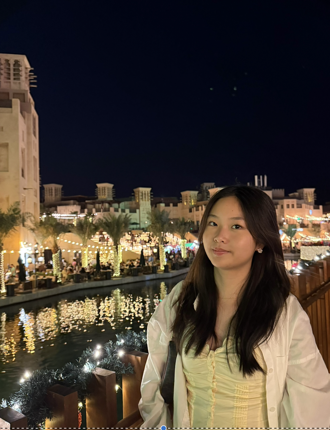A young woman with long dark hair standing near a canal at night, with city lights, palm trees wrapped in lights, and buildings in the background.