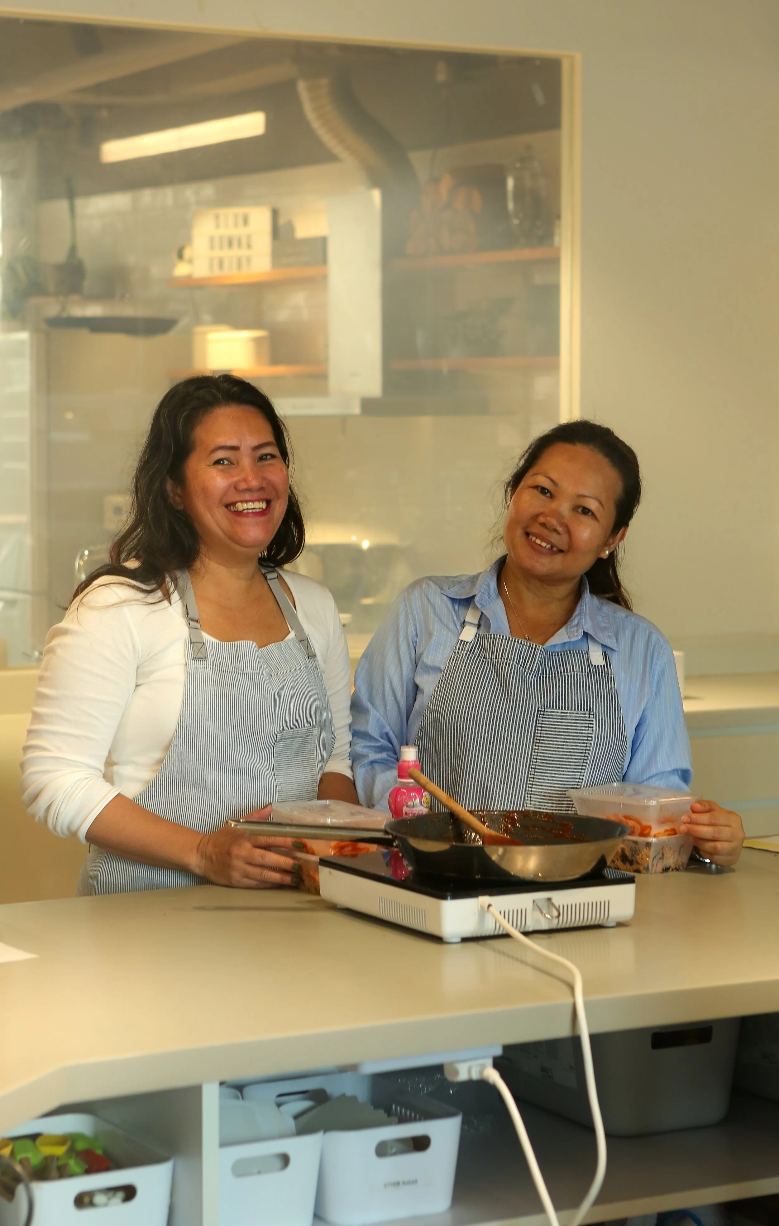 Two women wearing aprons standing in a kitchen, smiling at the camera, with a portable stove and cooking ingredients on the counter.