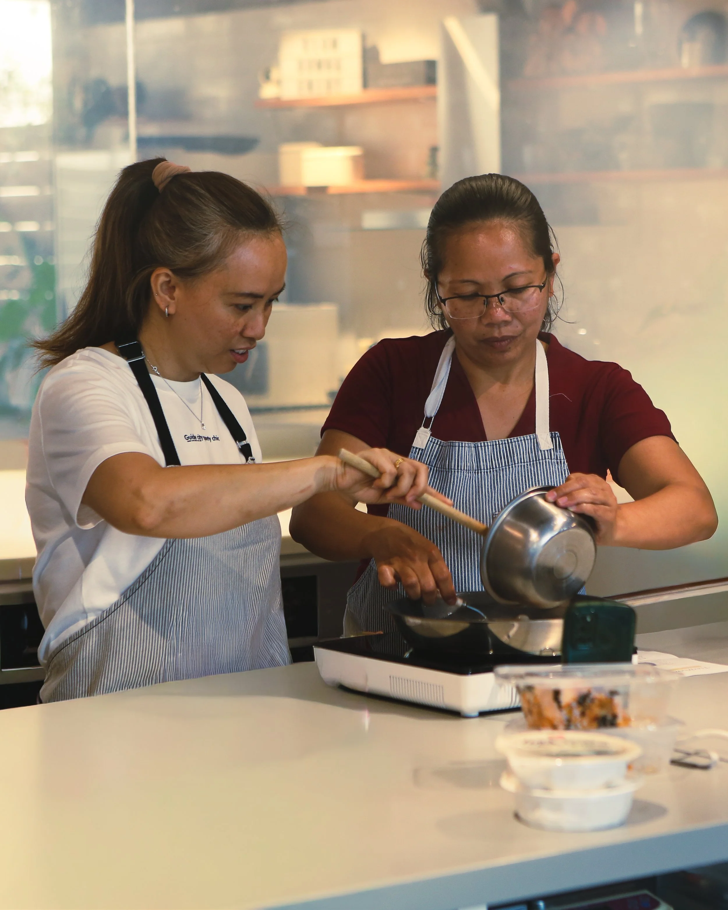 Two women cooking in a kitchen, one pouring batter into a pan, both wearing aprons.