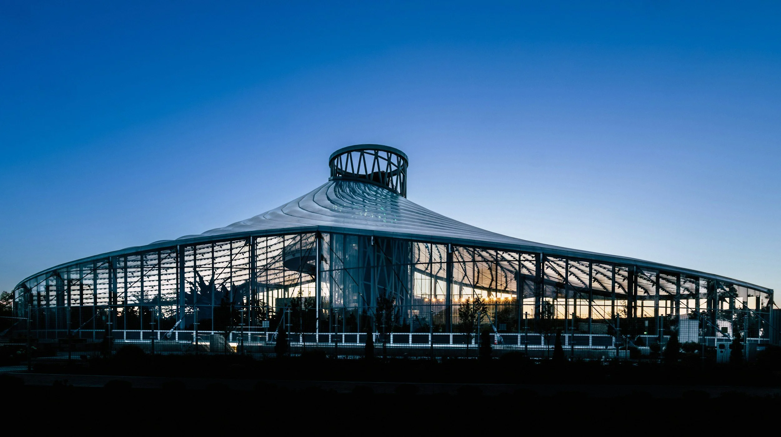 A modern glass greenhouse with a curved roof and a circular observation deck on top, illuminated by the setting sun, against a clear blue sky.