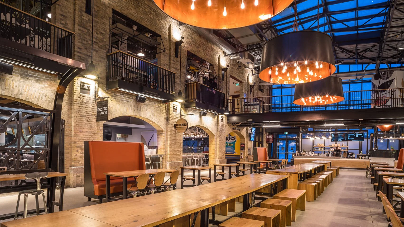 Empty modern restaurant interior with wooden tables, orange booth seating, black metal accents, brick walls, and large hanging black and copper light fixtures.