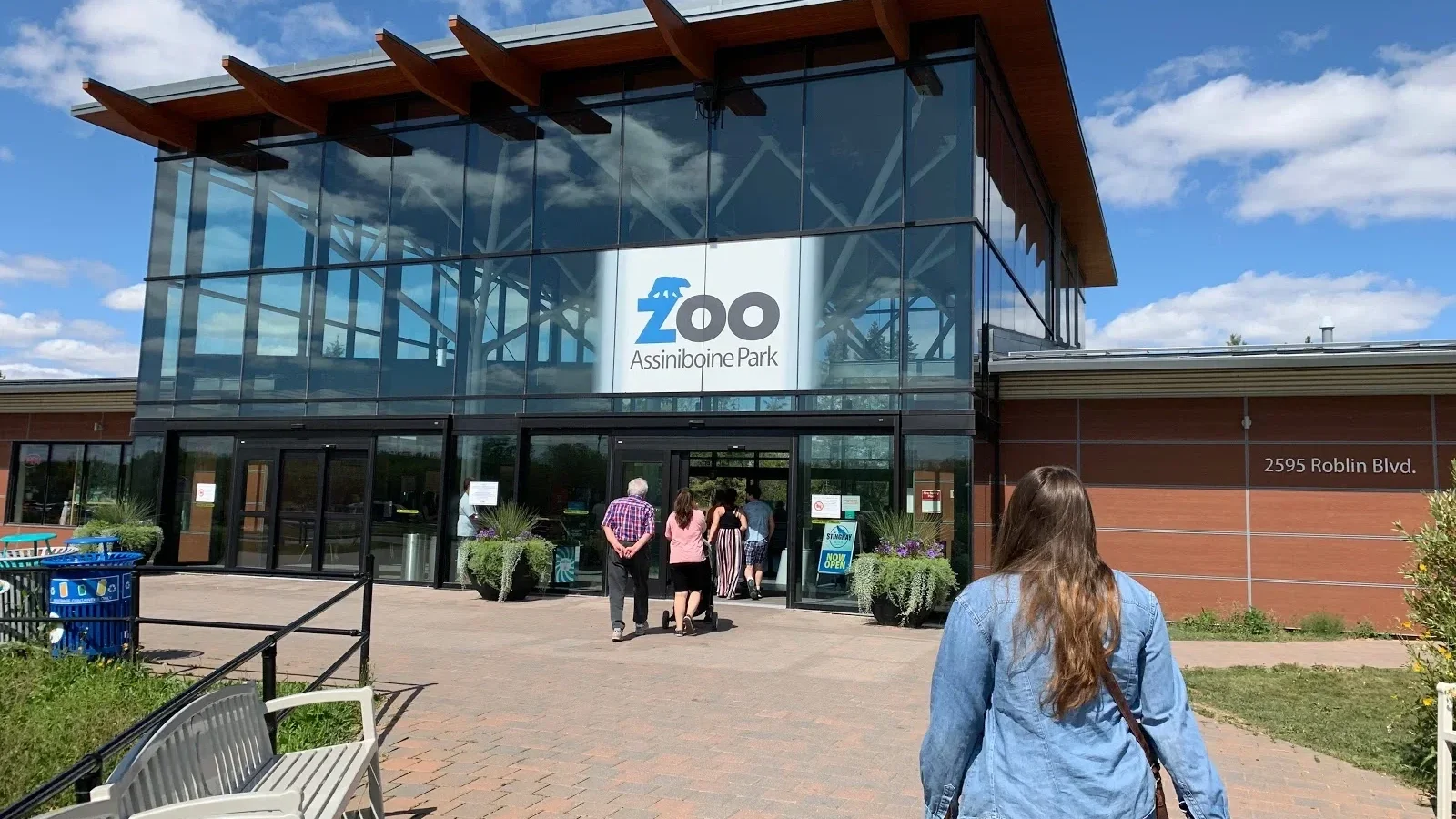 People entering Assiniboine Park Zoo with a sign that reads 'Zoo Assiniboine Park' on the building, which has a modern glass entrance, blue sky, and planters with flowers.
