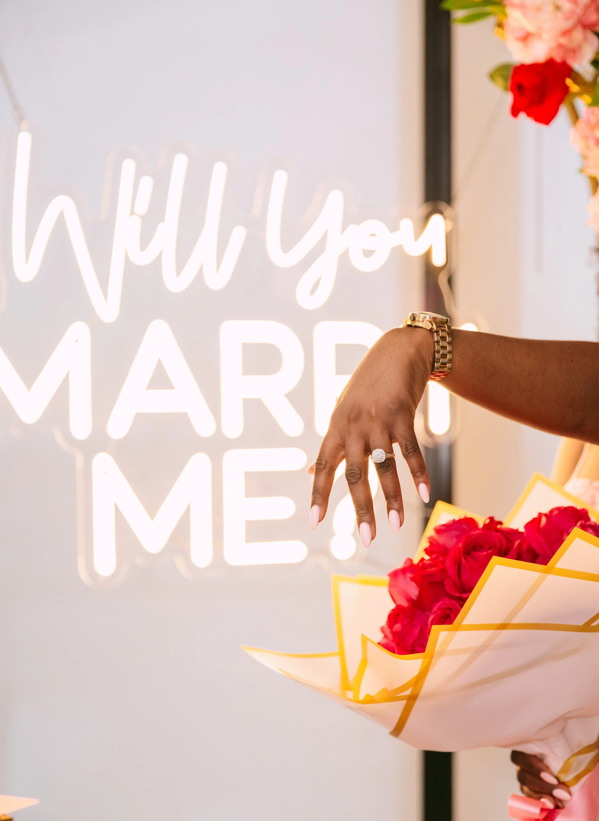 Close-up of a person's hand with a gold watch and diamond ring, holding a bouquet of pink and red roses, in front of a neon sign that reads 'Will you marry me?'