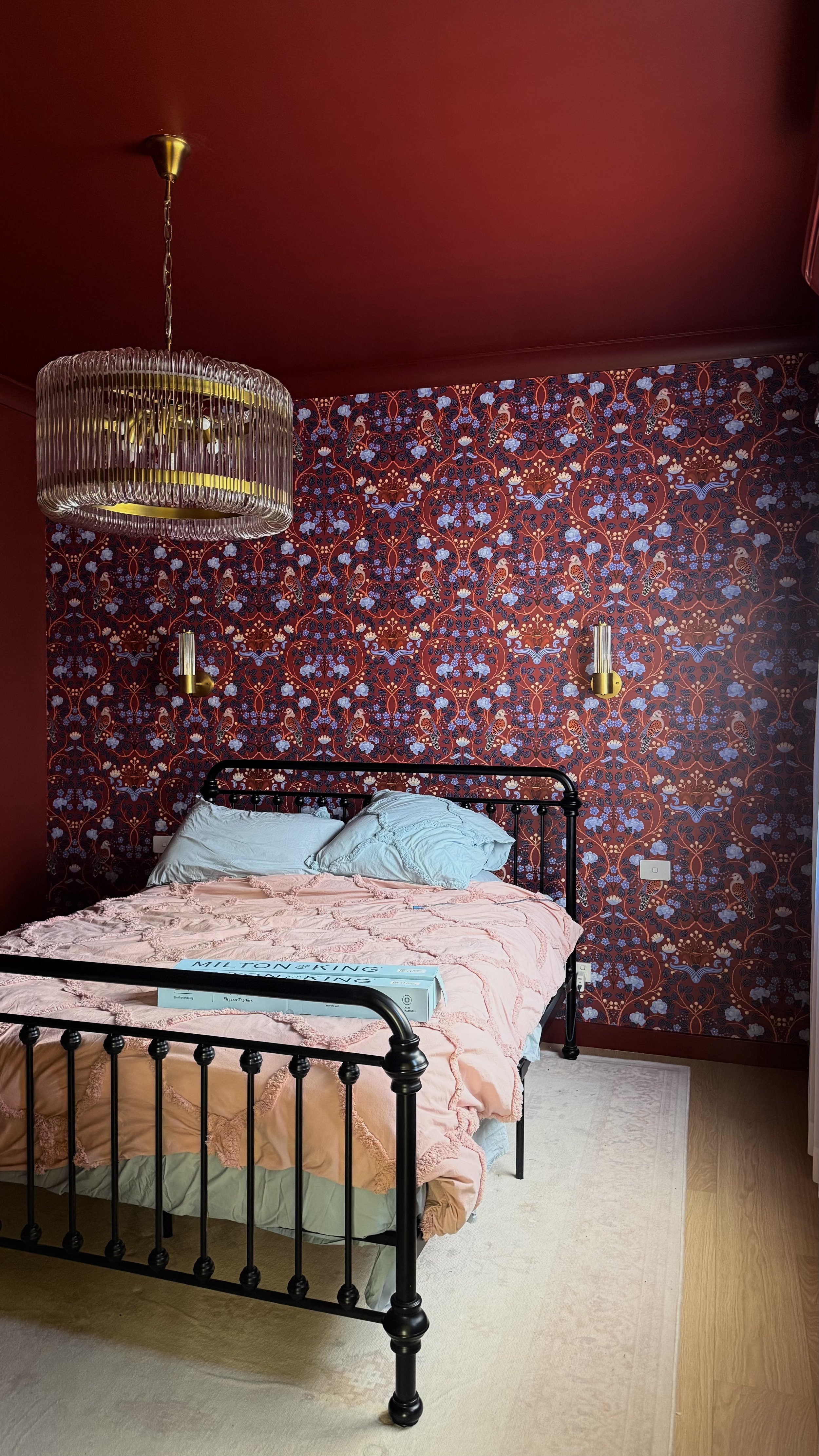 A bedroom with a black metal bed frame, pink bedding, and light blue pillows. The wall behind the bed has a red floral wallpaper. There are two gold wall sconces and a modern hanging light fixture. A box of Milton & King wallpaper is on the bed.