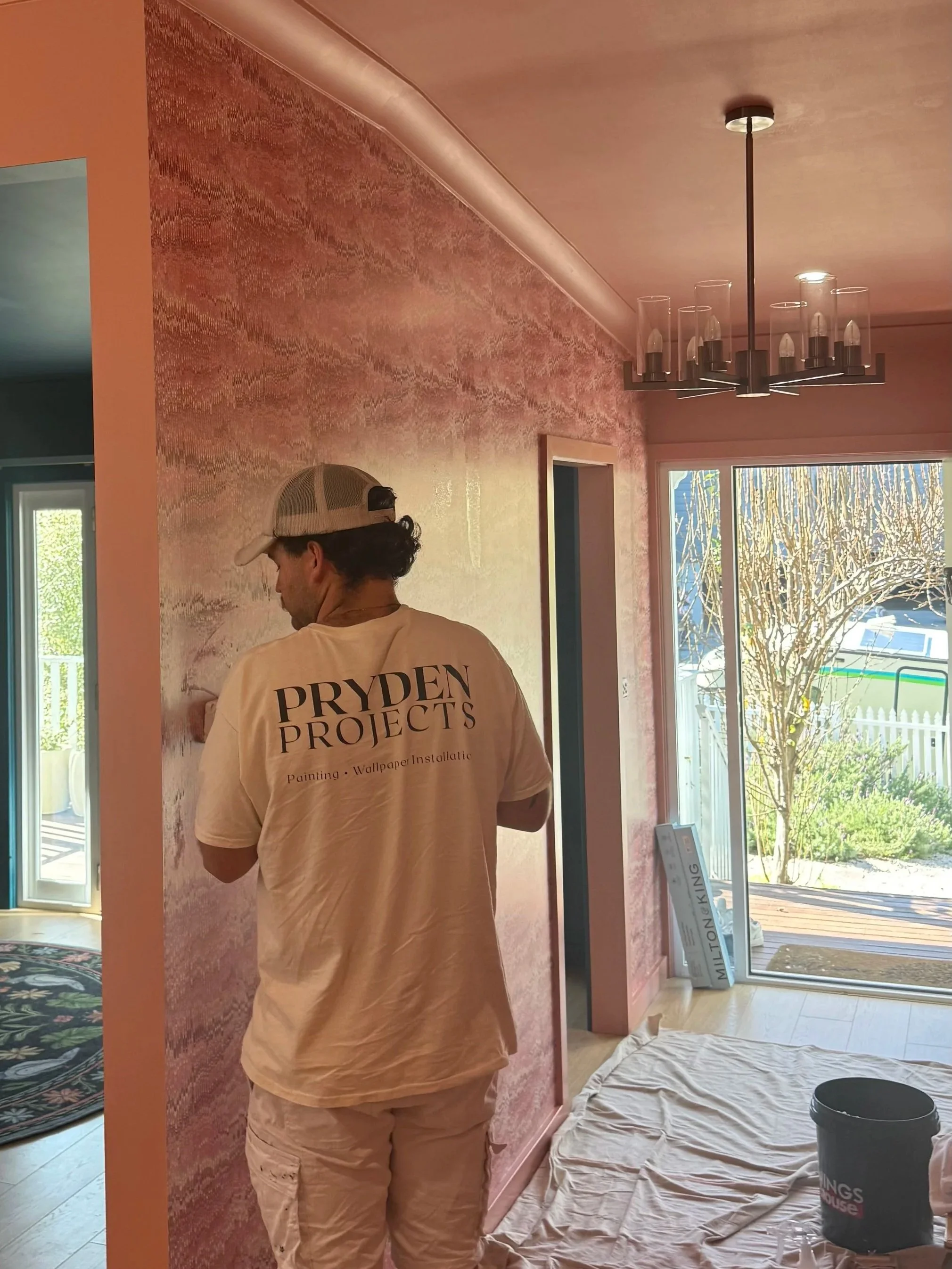 A person wearing a beige Pryden Projects shirt, beige pants, and a beige baseball cap is painting a wall pink with a textured finish. The room has large window sliding doors letting in natural light, with trees and a yard visible outside. Painting supplies, including a bucket and a plastic sheet on the floor, are present.