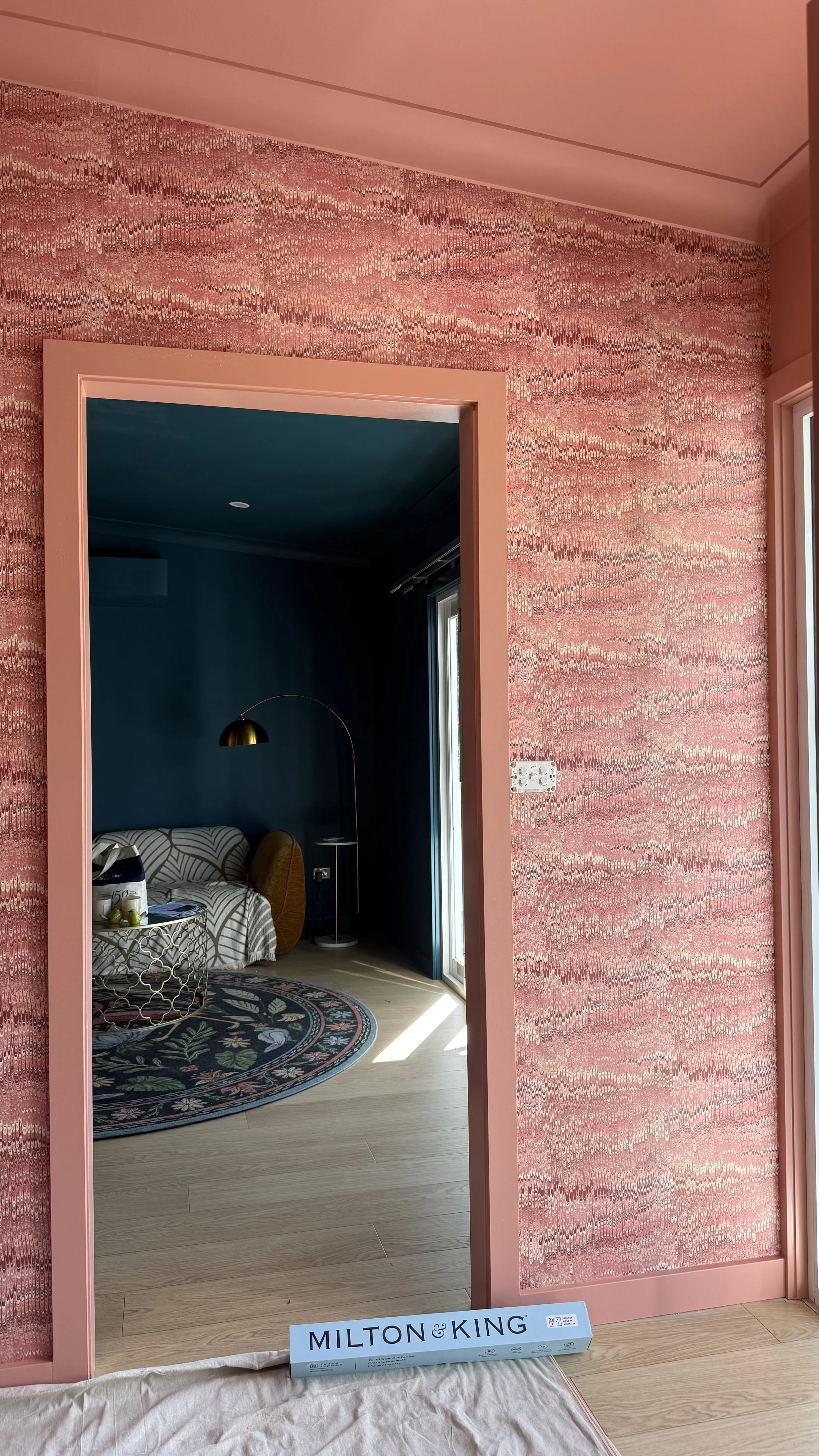 Interior of a room with pink textured wallpaper, door frame, and a view into a dark blue room with a patterned rug, sofa, and standing lamp.