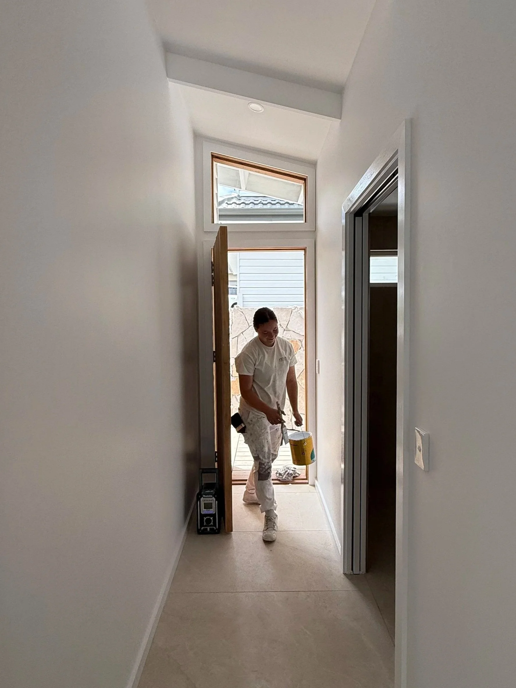 Person in a white shirt and pants holding a bucket, walking through a narrow hallway toward an open door with a view outside to a tiled roof and sky.