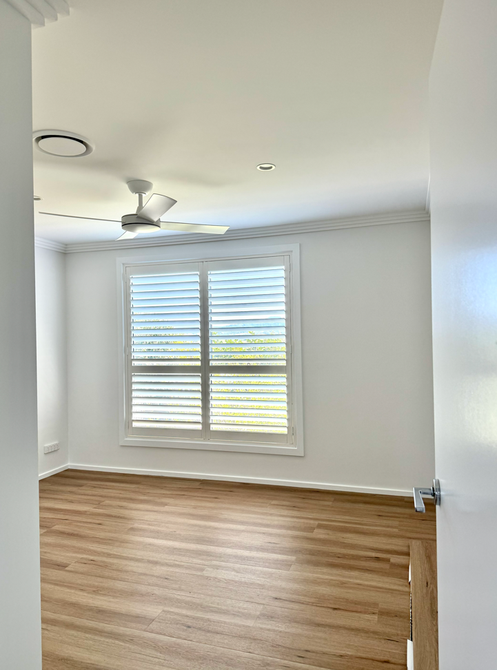 Empty room with white walls, a large window with white shutters, a ceiling fan, and wooden flooring.