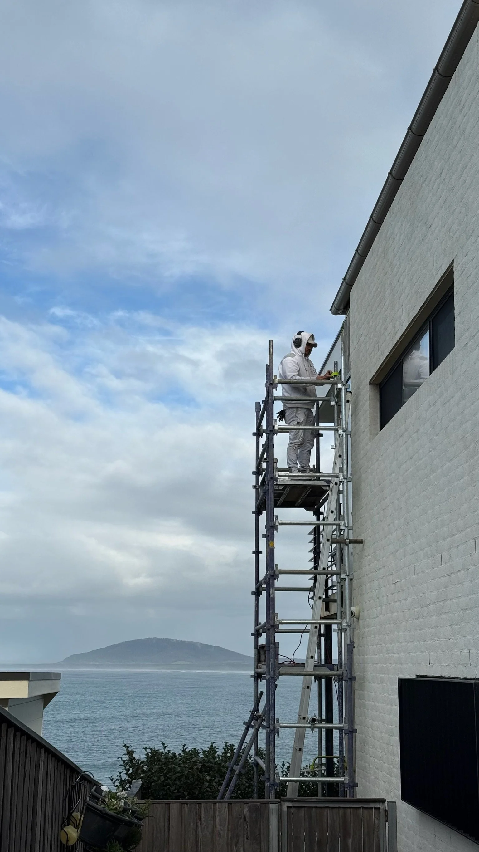 A construction worker wearing a white outfit, gloves, and headphones standing on scaffolding next to a white brick building, with a body of water and an island in the background.