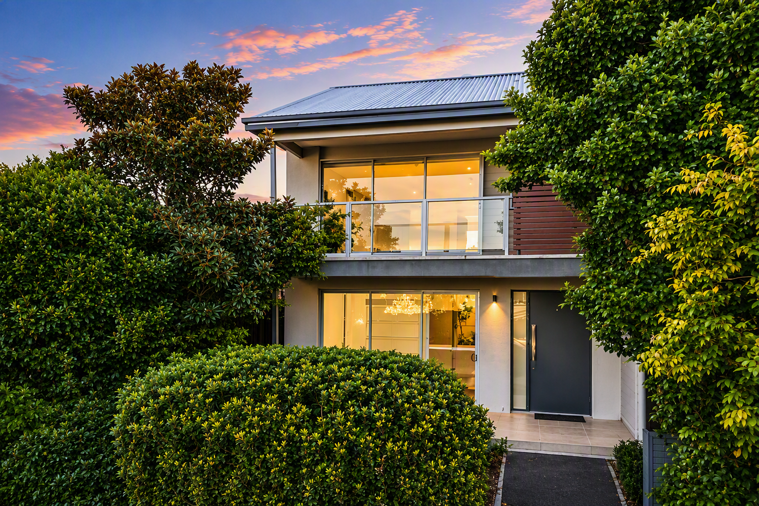 Modern two-story house with large windows, surrounded by lush green bushes and trees, at sunset.