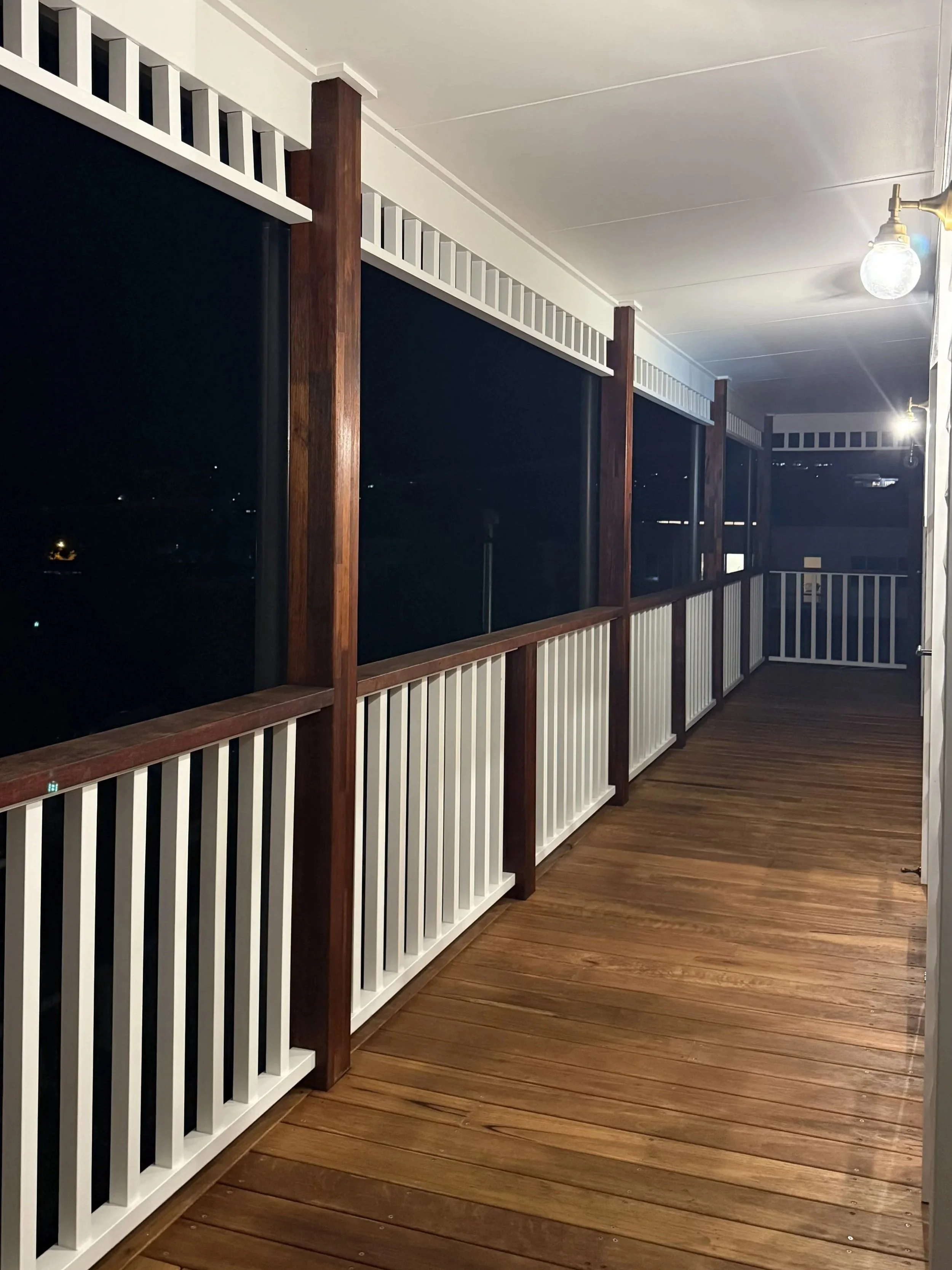 A covered balcony or porch area at night with white and brown wooden railing, wooden floorboards, and a ceiling-mounted light bulb.