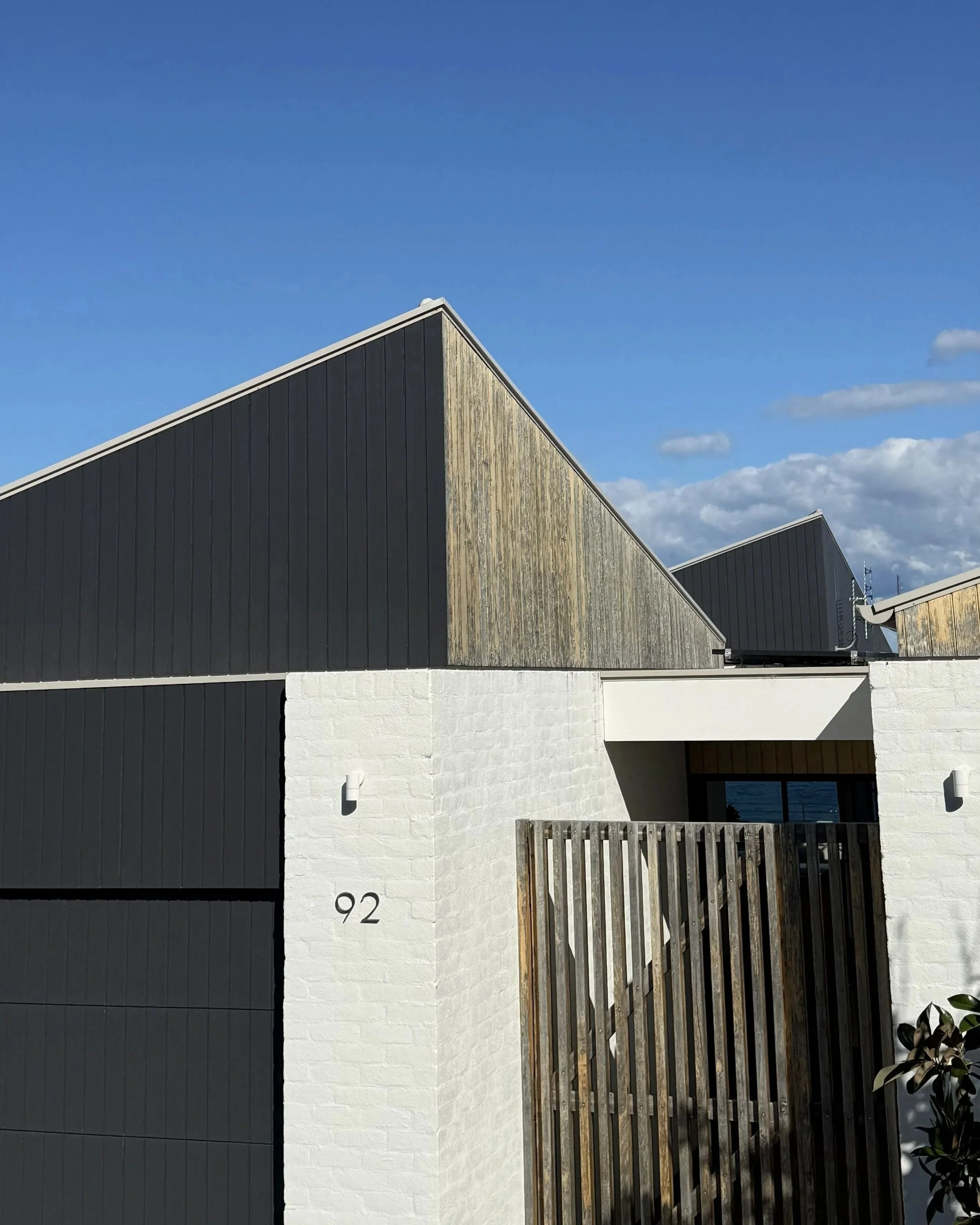 The image shows a modern building with black vertical panels and a white brick section with the number 92 on it, with a wooden fence in front. The sky is blue with some clouds.