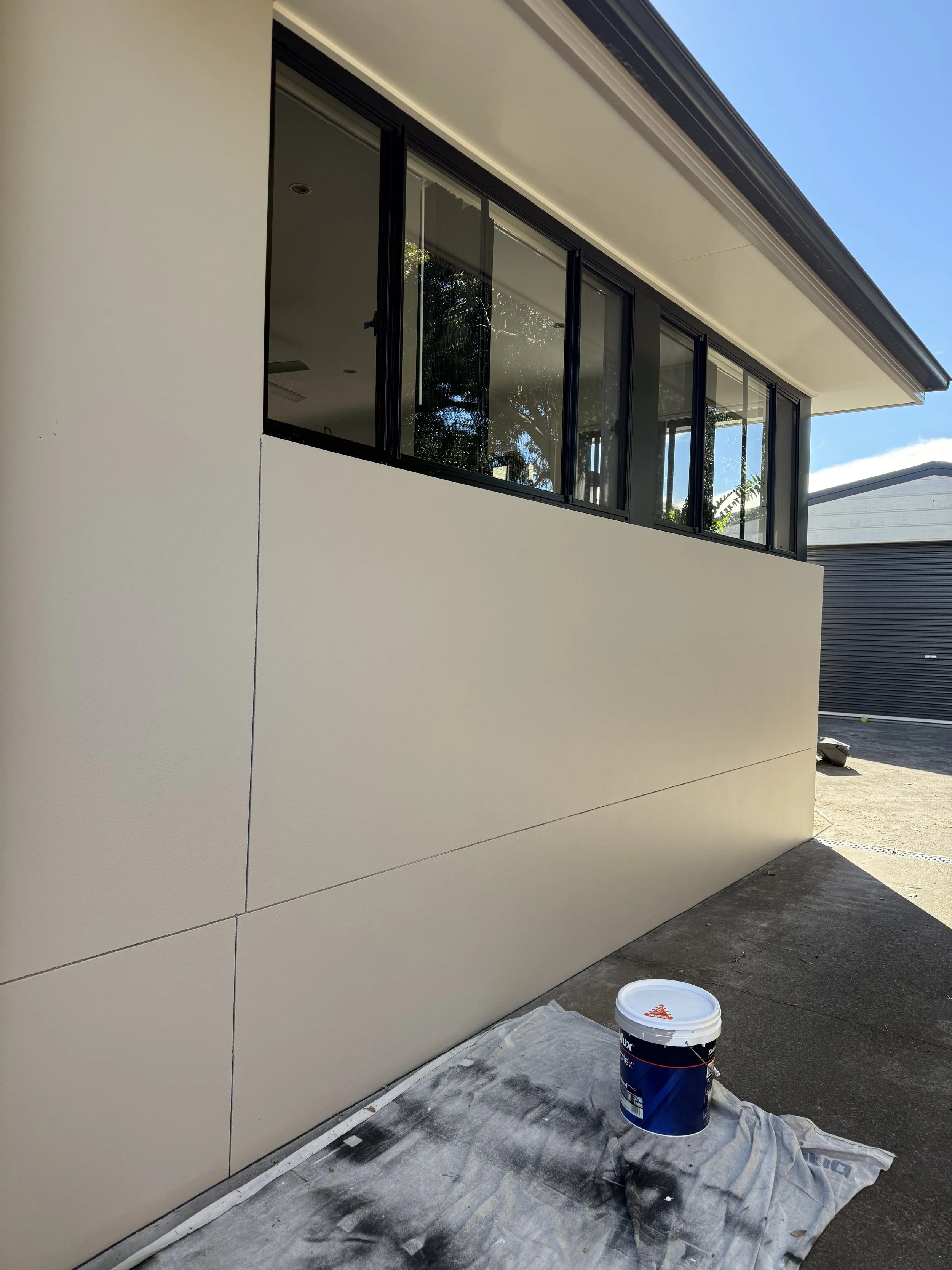 Exterior of a modern house under construction or renovation, showing a large white wall and a section of black-framed glass windows, with painting supplies on the ground.