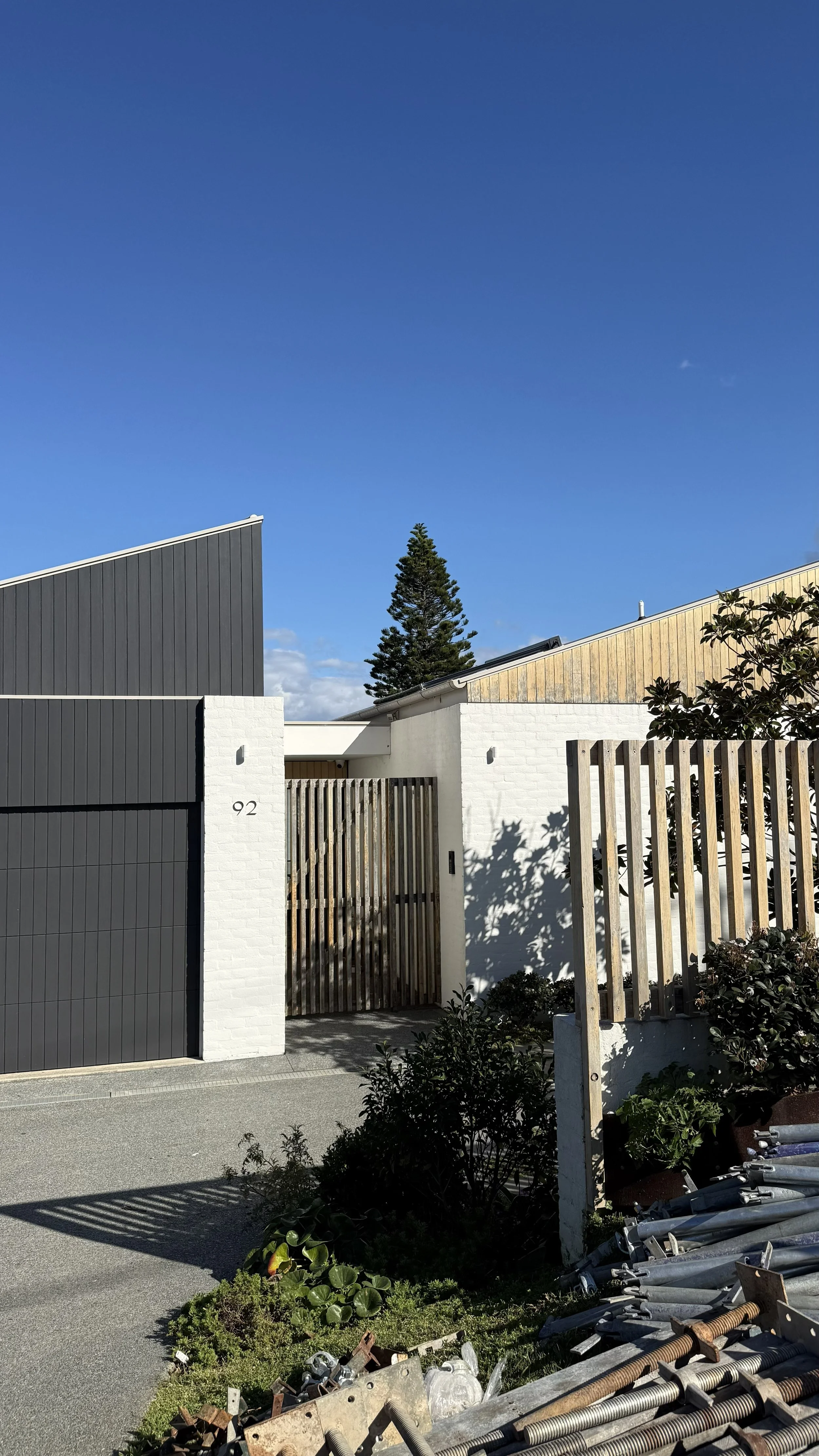 A modern house with a black garage door and a white brick wall, a wooden gate, and landscaping with plants and shrubs. There are construction or industrial materials in the foreground.