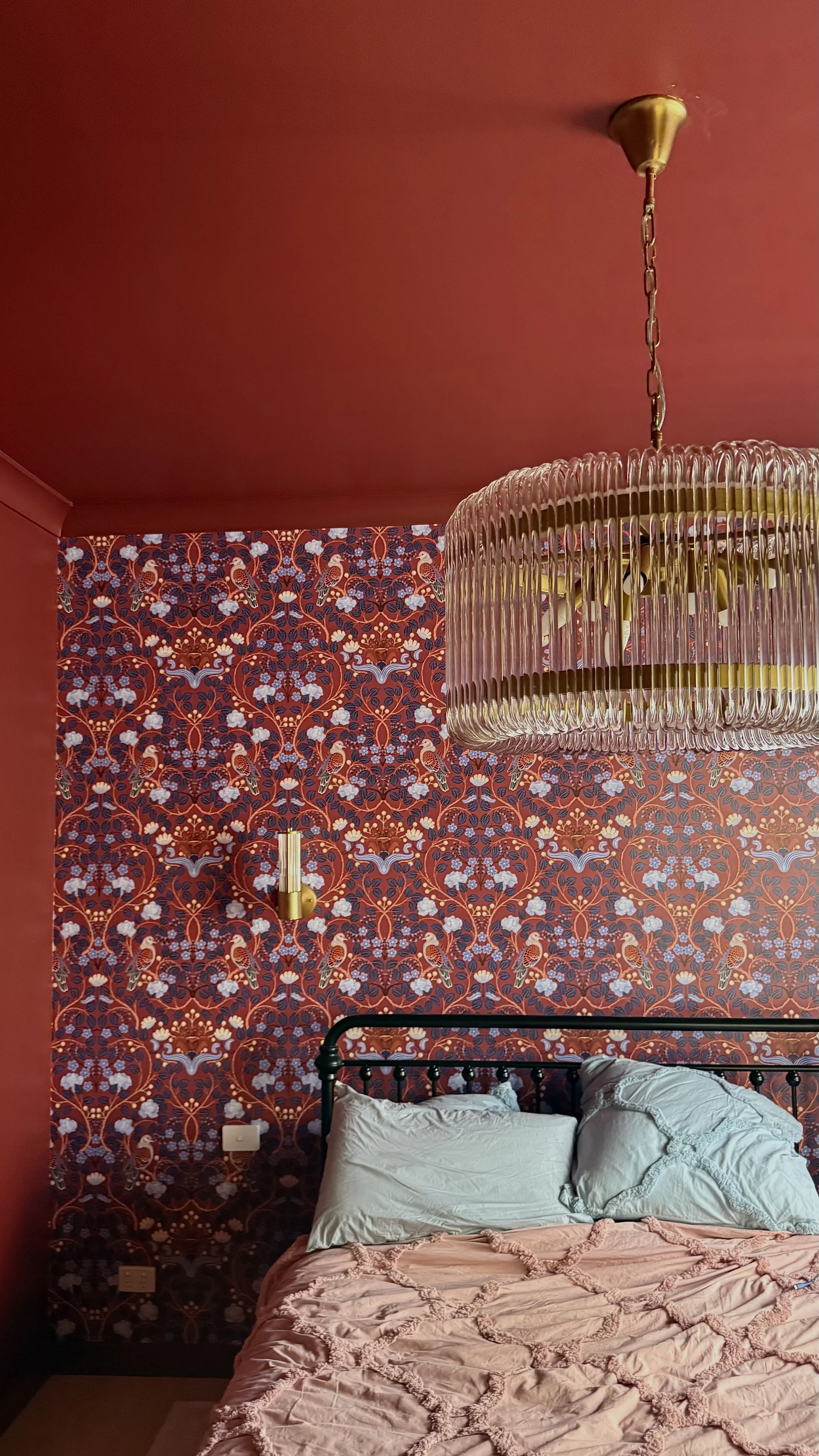 A bedroom with a red and patterned accent wall, a black metal bed with white pillows, a pink quilt, a gold wall sconce, a large glass chandelier, and a red ceiling.