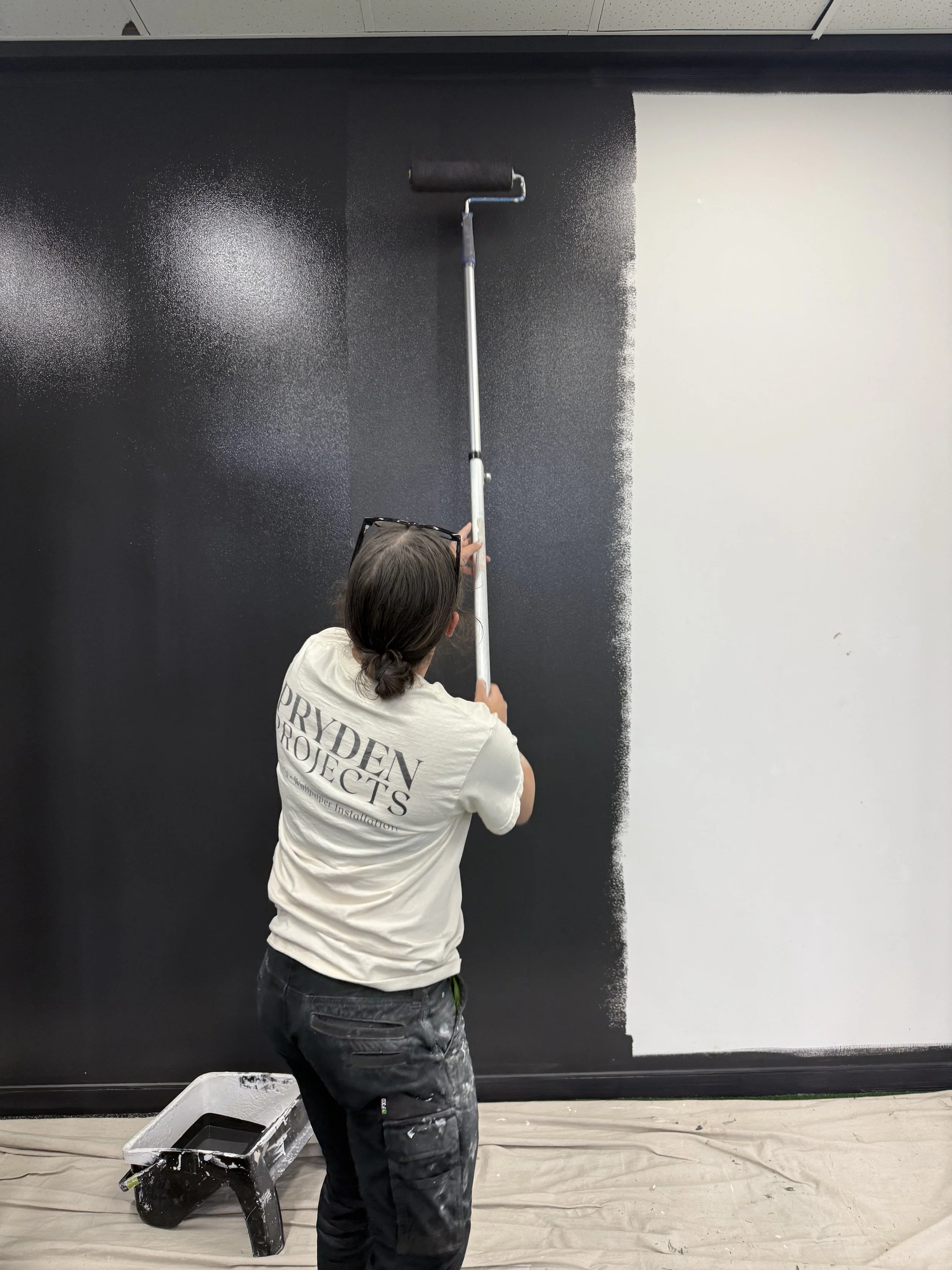 Person spray painting a large wall with black paint, wearing a shirt that says 'Princeton Projects,' with a paint roller, a paint tray, and a drop cloth on the floor.