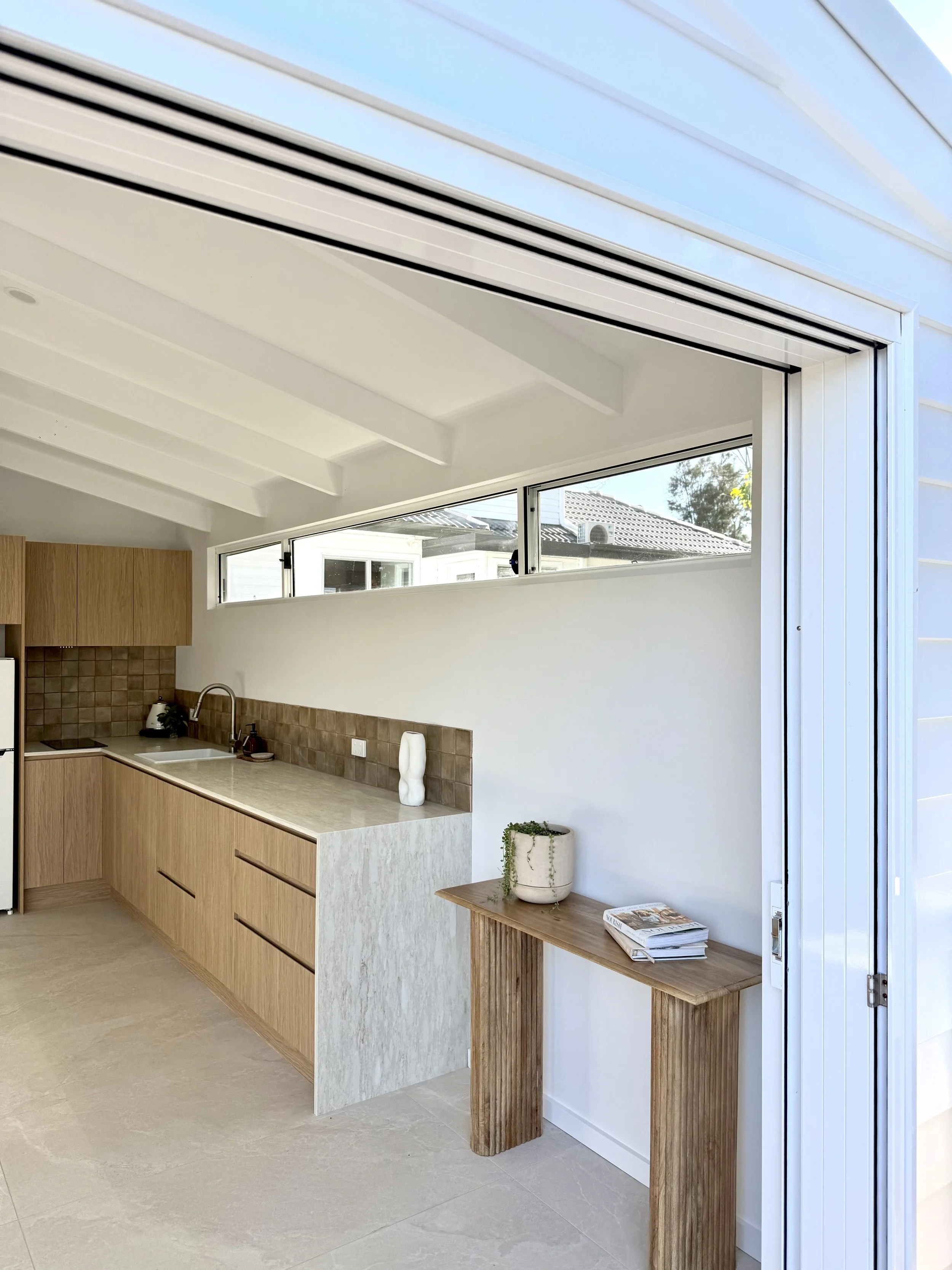 Kitchen with beige cabinets, a beige countertop, a small sink, and a window with a view of a neighboring house and trees.