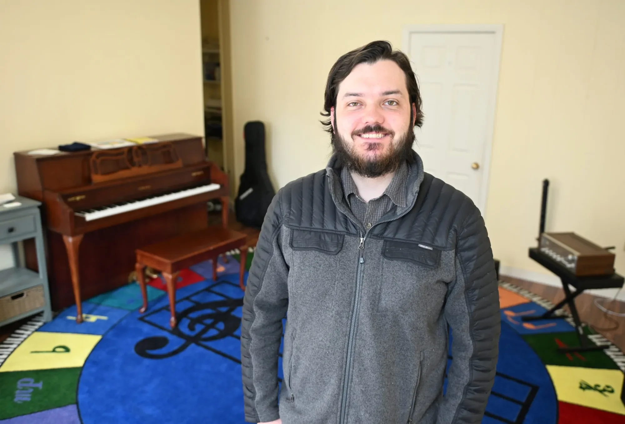A smiling man with dark hair and beard wearing a gray jacket standing in a room with a piano, colorful music symbol rug, and various musical instruments.