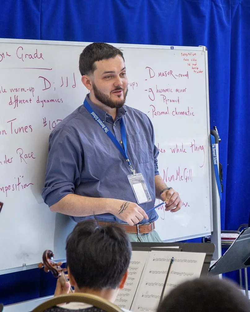 A music teacher standing in front of a whiteboard in a classroom, holding a baton, with students playing string instruments in the foreground.