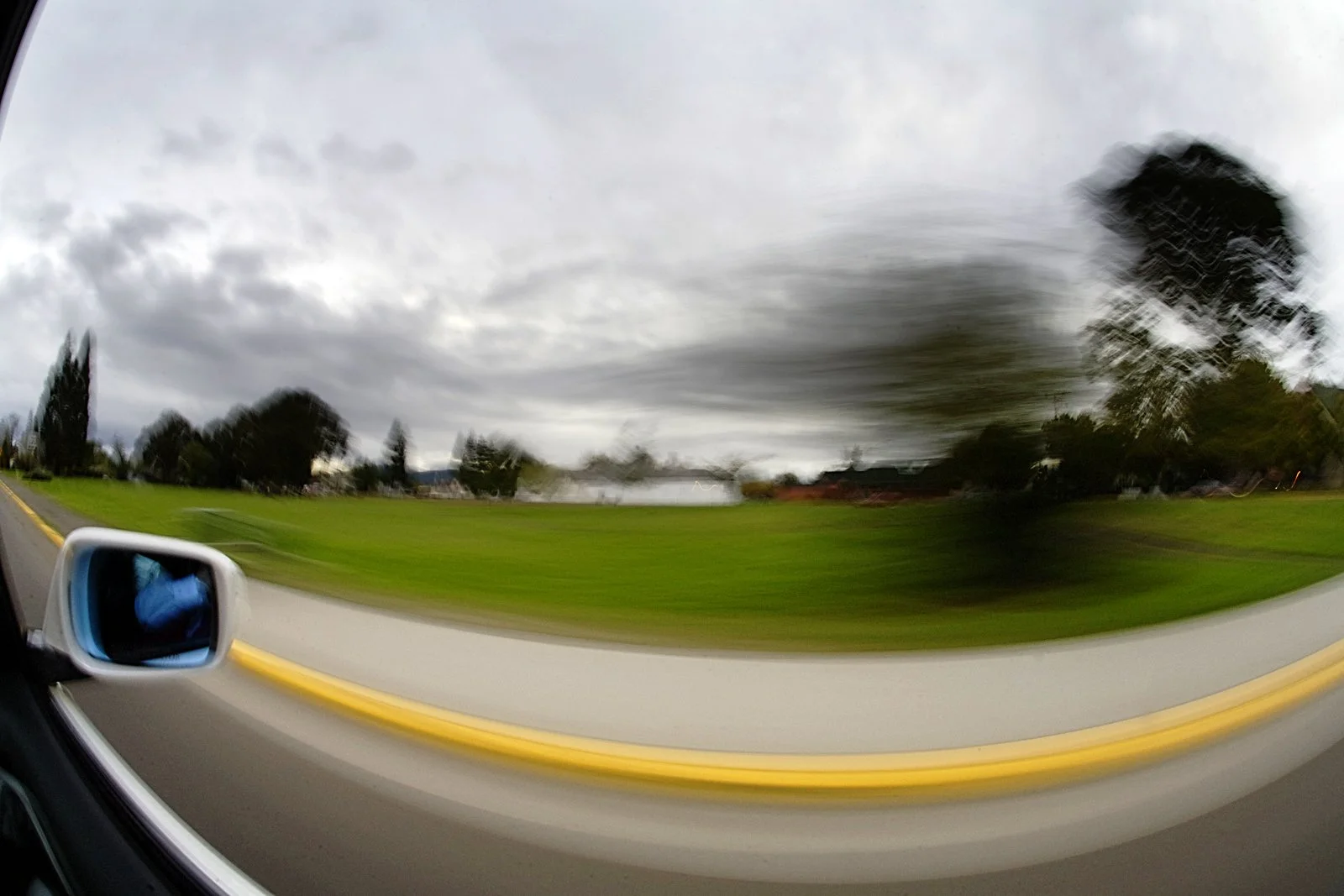 Motion blurred view from inside a moving vehicle showing a side mirror, a road, green grass, trees, and a cloudy sky.