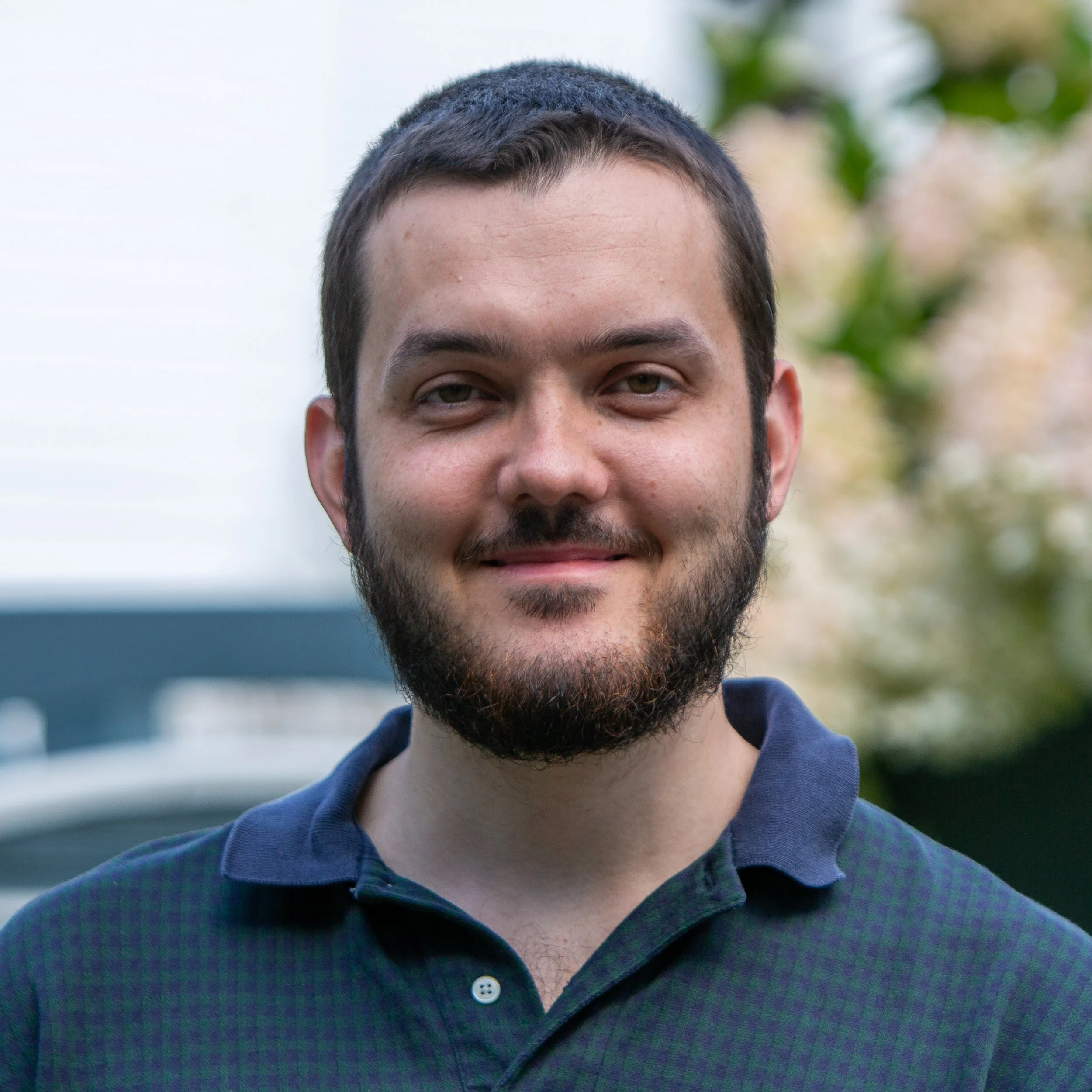 A young man with short dark hair and a beard, smiling outdoors with blurred greenery in the background.