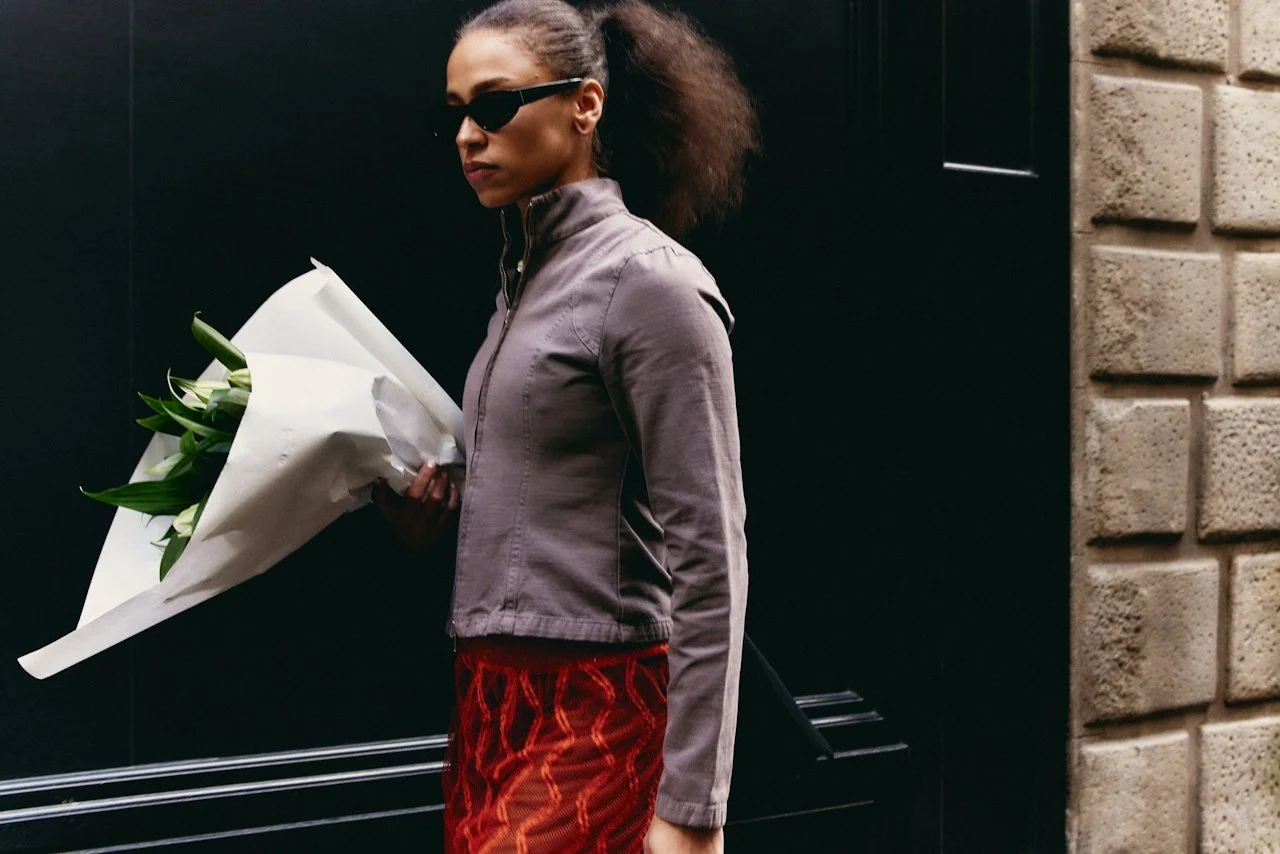 A woman with curly hair pulled back, wearing dark sunglasses and a gray jacket, holding a bouquet of white flowers wrapped in white paper, standing against a black and beige brick wall.