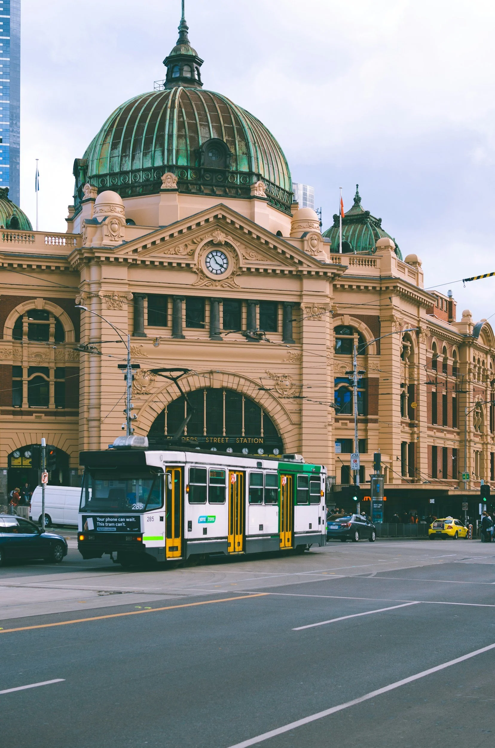An old-fashioned beige building with a dome and clock, and a white tram with yellow details in front of it on a city street.