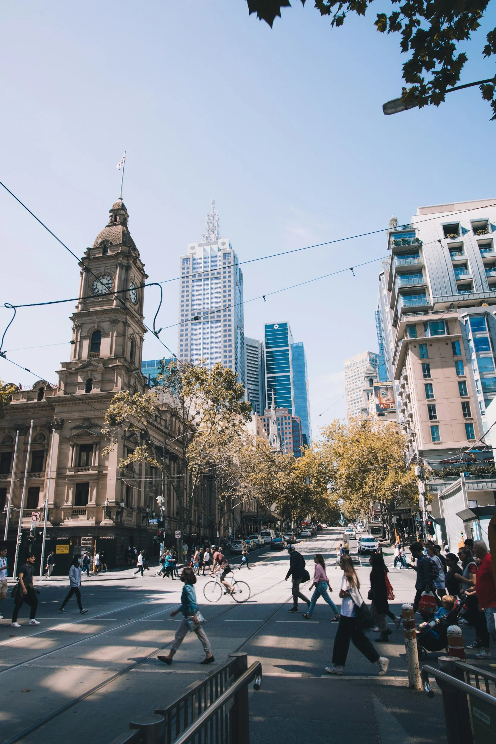 City street with pedestrians crossing, tall skyscrapers, and a historic building with a clock tower under a clear blue sky.