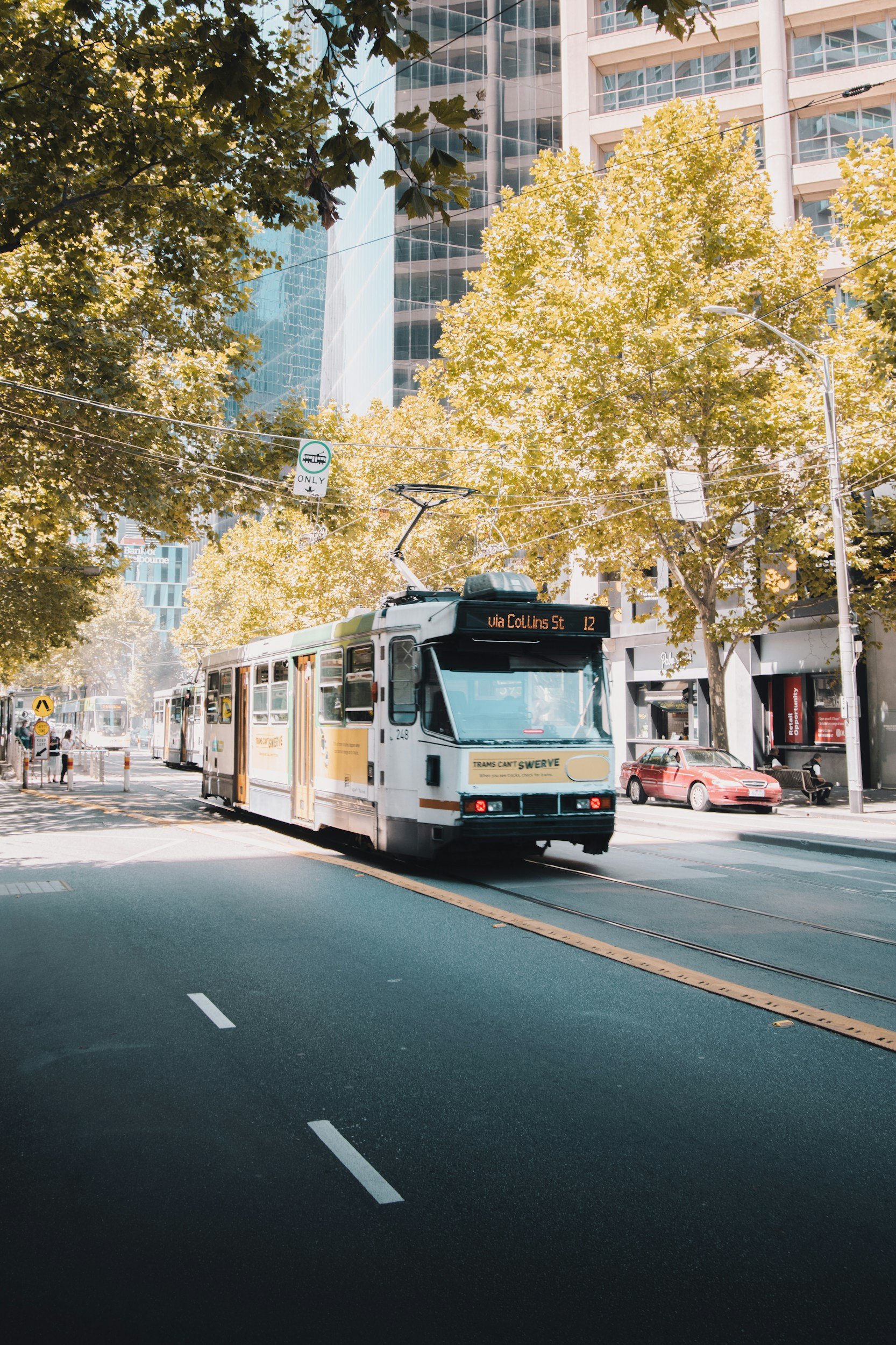 A city street with a tram in the center, trees with green and yellow leaves, and modern glass buildings in the background.