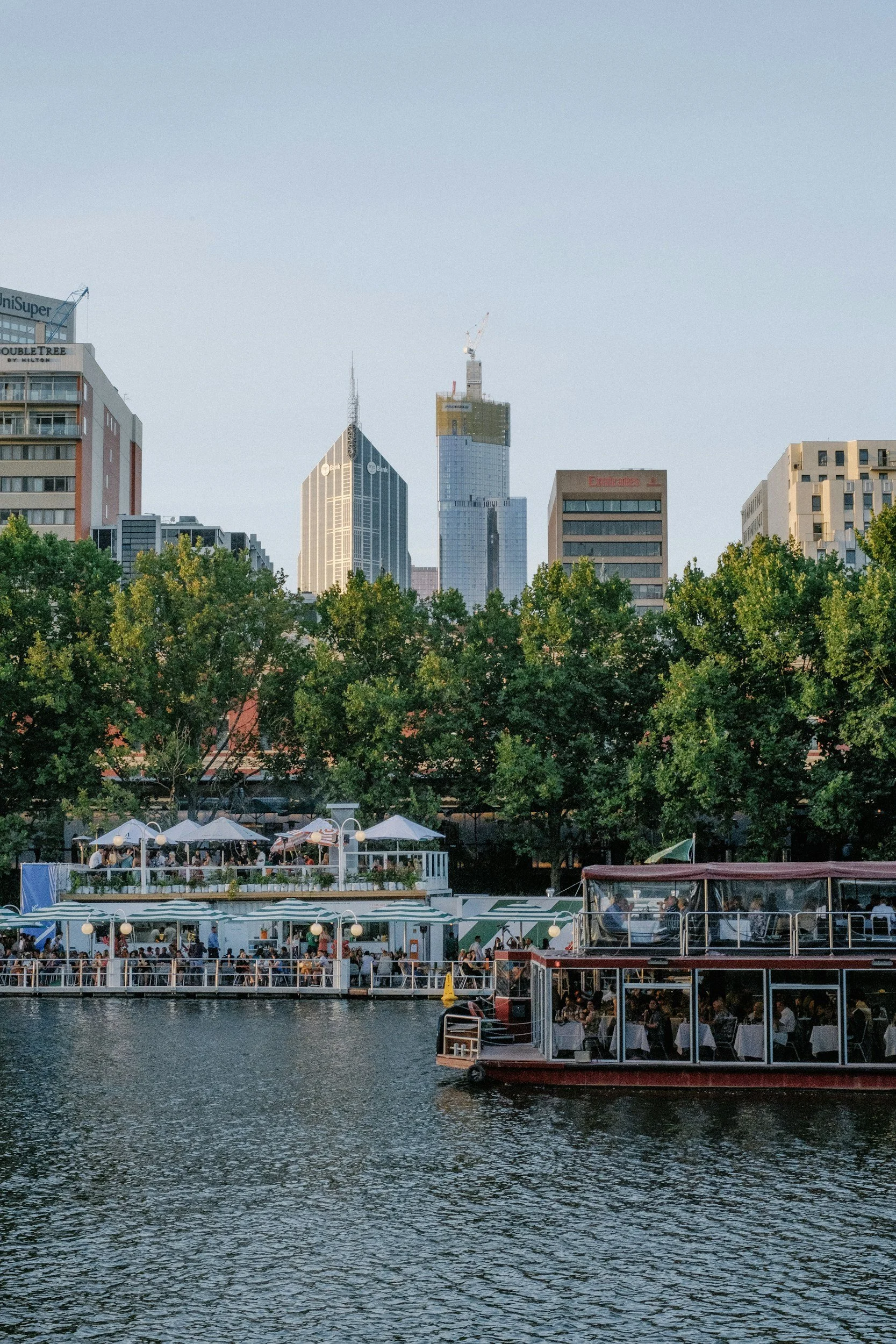 City skyline with tall buildings, trees, and a river with boats and restaurant patios along the water.