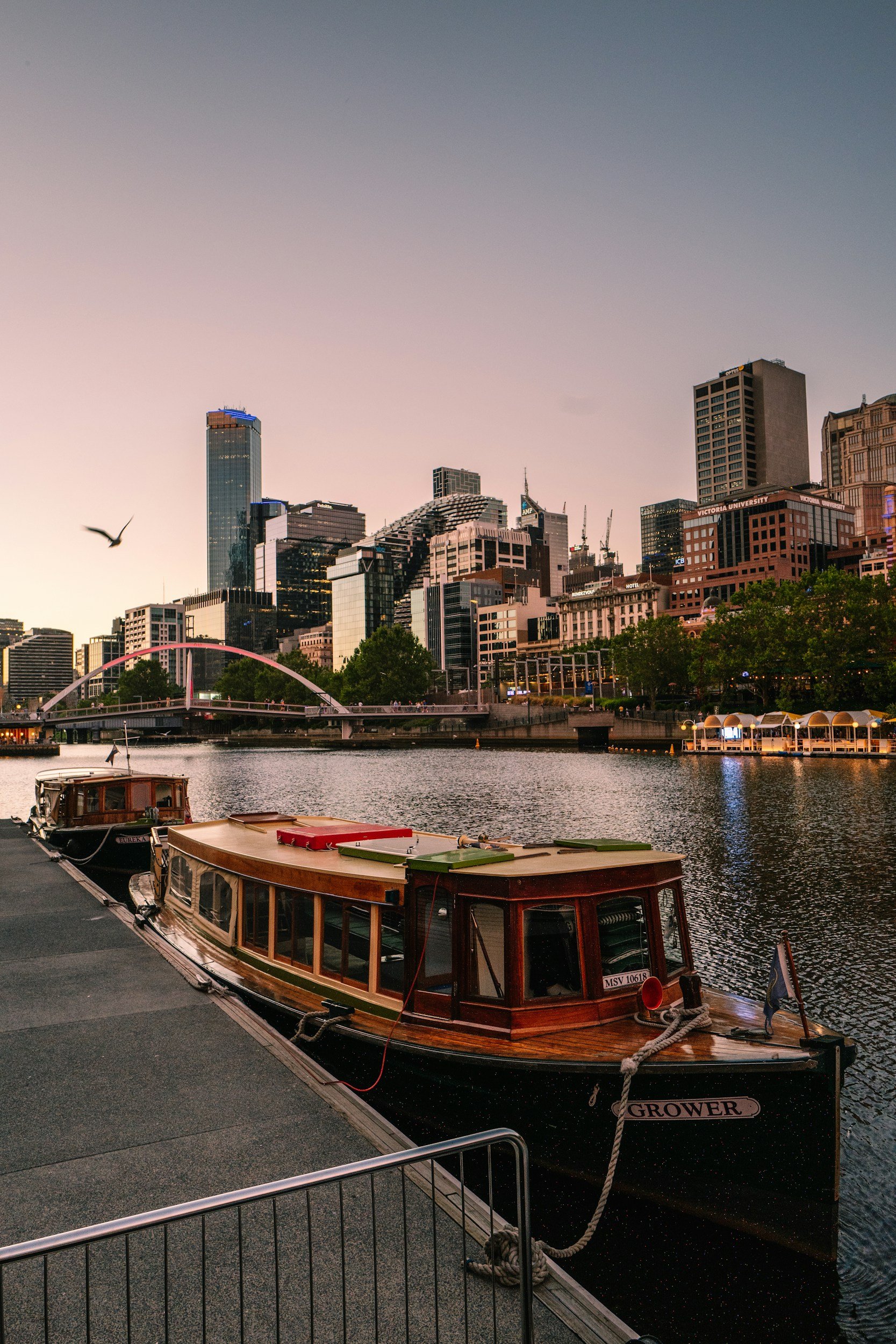 A wooden boat named 'Grower' docked along a waterfront with a city skyline in the background during dusk or dawn.