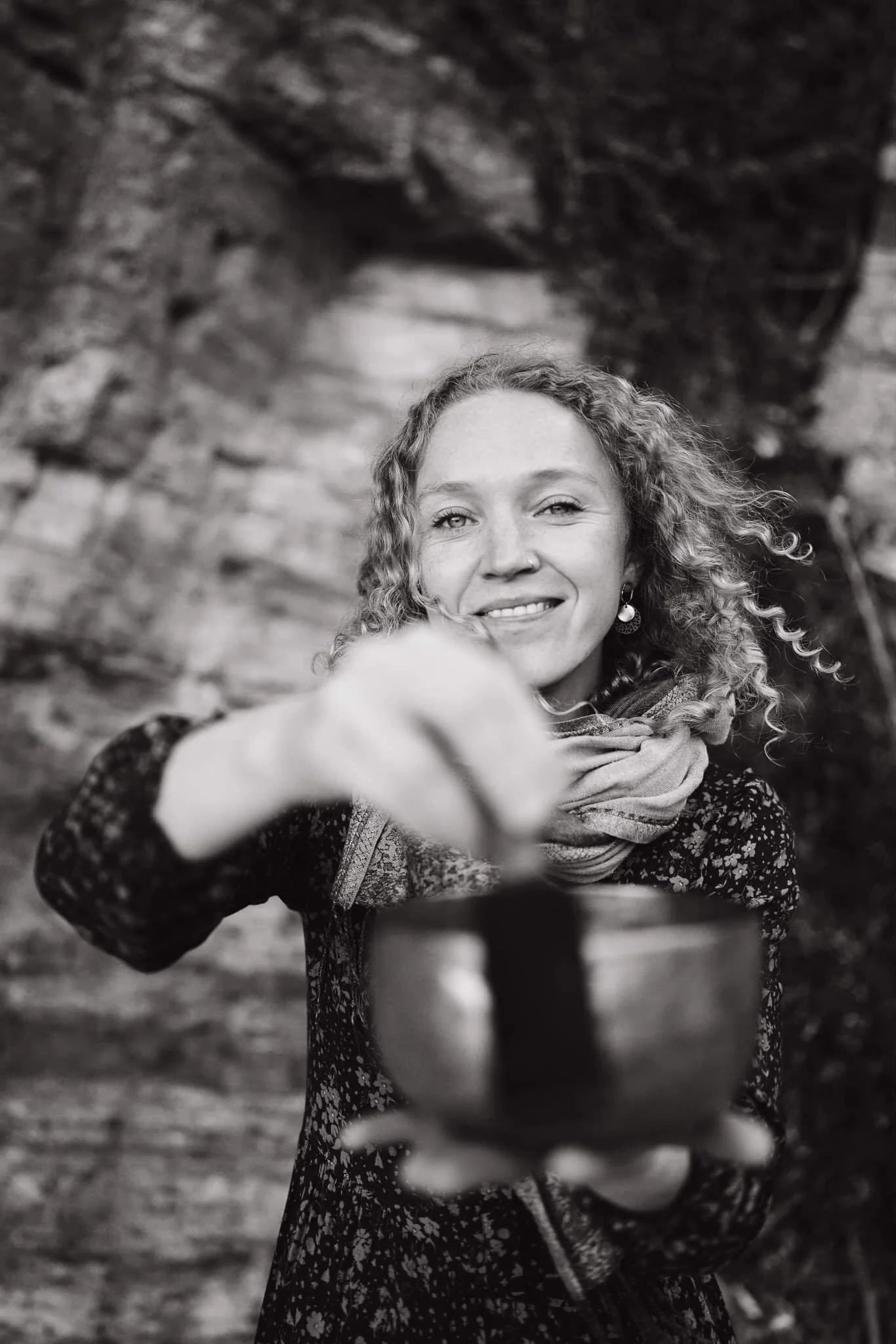 A woman with curly hair smiling and holding a bowl, pointing towards the camera, standing outdoors with a rocky background.