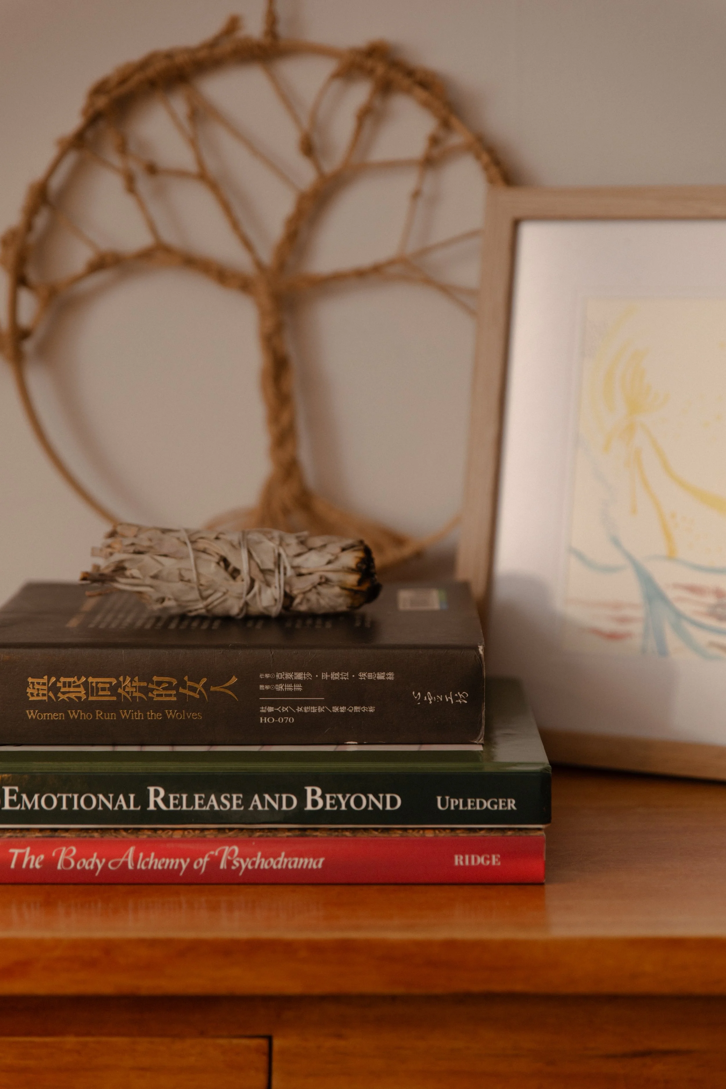A wooden table with three stacked books, a bundle of sage, and a framed artwork. In the background, a decorative rattan tree wall hanging.