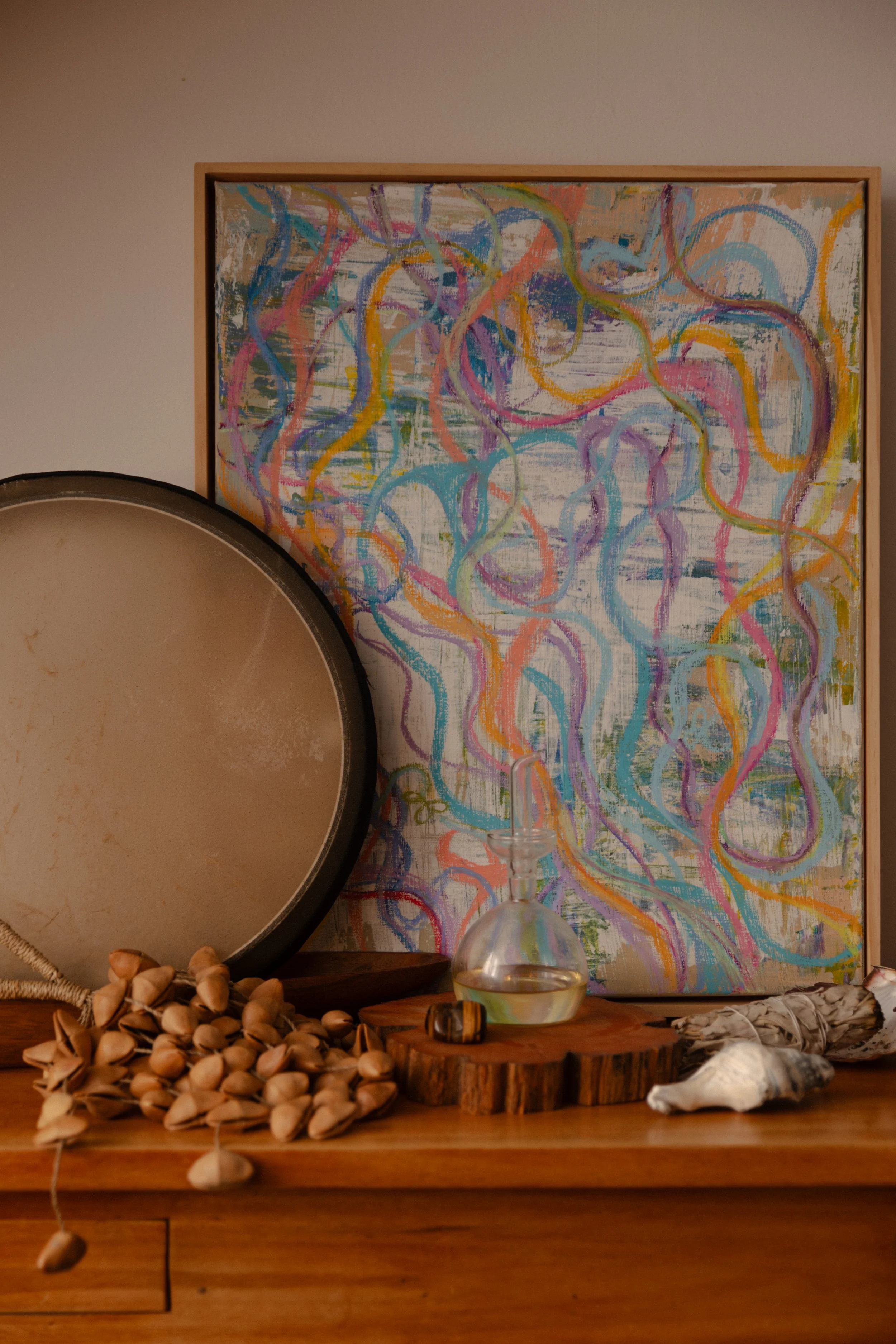 Decorative scene with an abstract painting with colorful squiggly lines, a large drum, a small glass decanter with water, dried pistachio shells, and seashells on a wooden surface.
