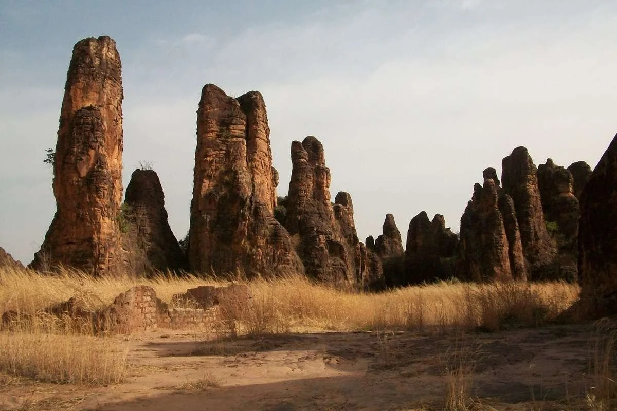 A landscape of tall, reddish-brown rock formations in a desert area with dry yellow grass in the foreground and a mostly cloudy sky in the background.