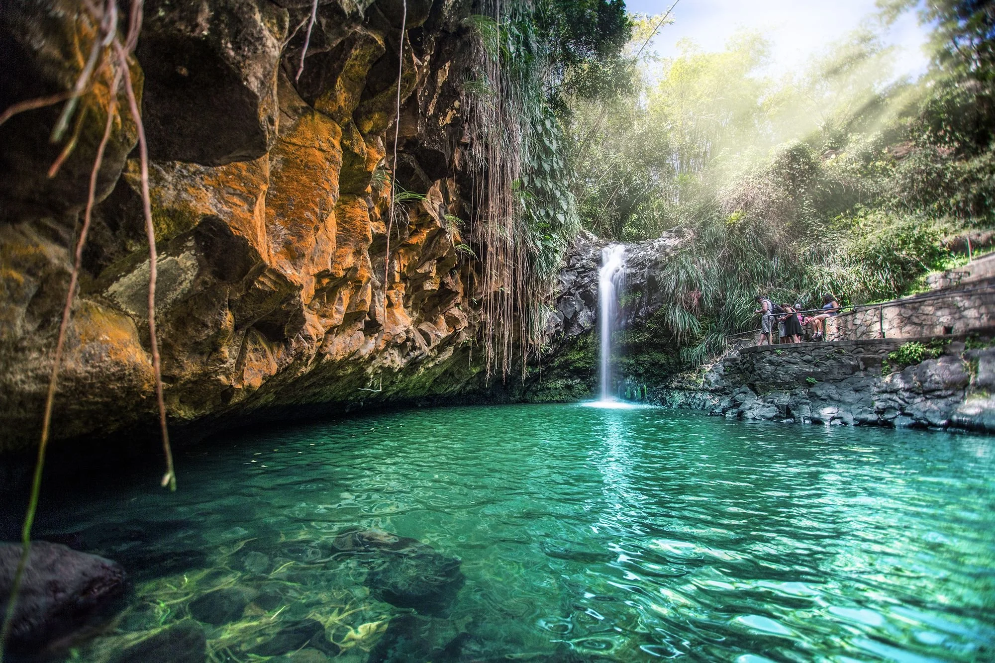 A waterfall cascading into a clear, greenish pool surrounded by rocky terrain and lush vegetation, with a group of people standing on a pathway in the background.