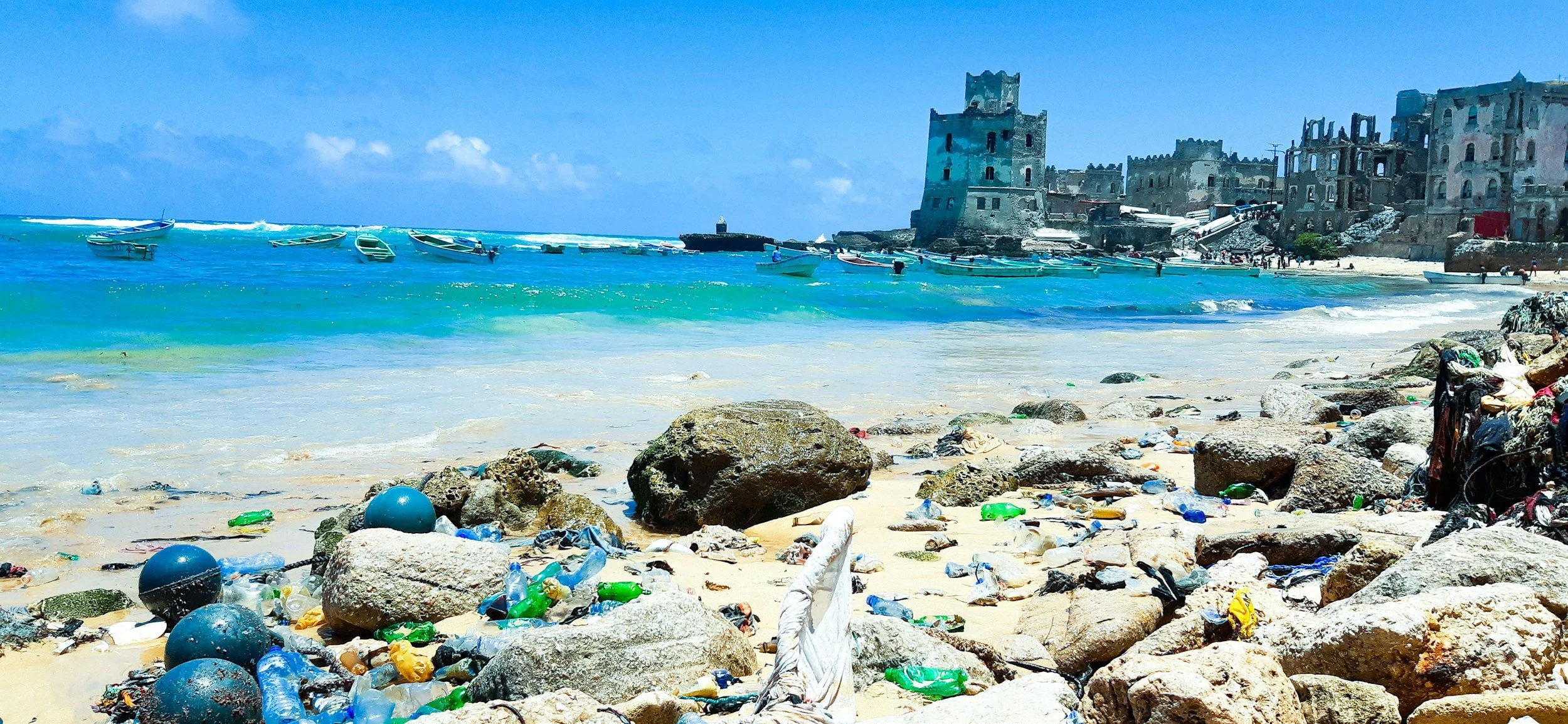 A beach scene with clear blue water, boats anchored near the shore, and an old ruined building on a hill in the background. The sandy beach is littered with rocks, debris, and trash.
