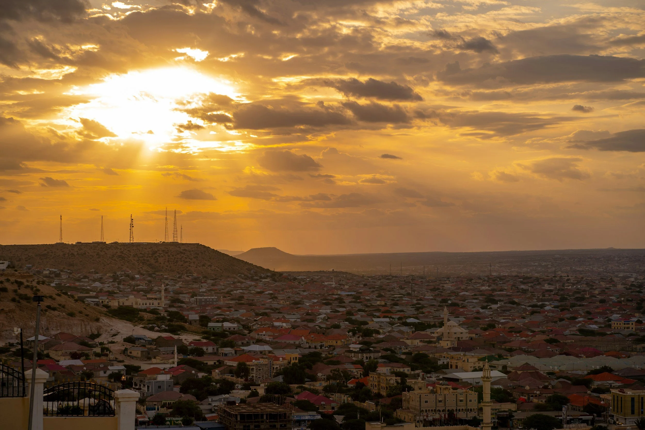 Sunset over a city with houses, hills, and transmission towers.