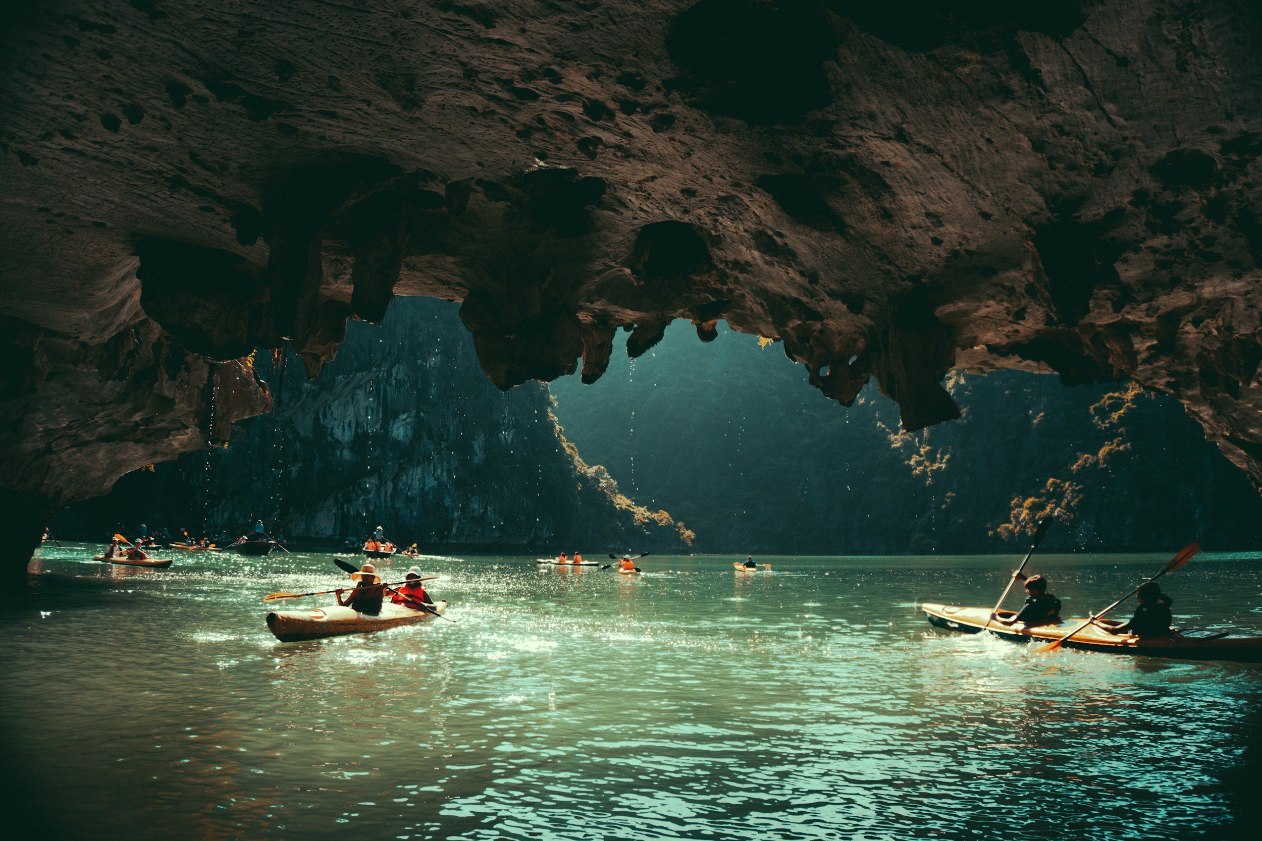People kayaking inside a natural cave with sunlight shining through opening and water surrounding them.