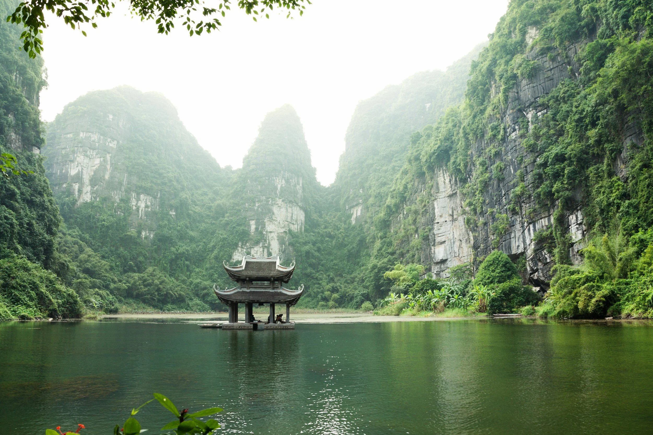 A serene landscape of a river with a traditional Asian pavilion in the center, surrounded by lush green mountains and trees.