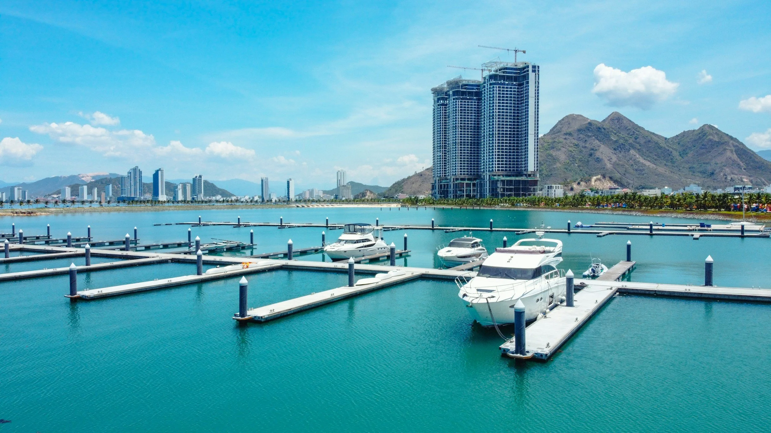 Marina with multiple yachts docked at floating piers, city skyline with tall buildings, mountains in the background, and blue sky with scattered clouds.