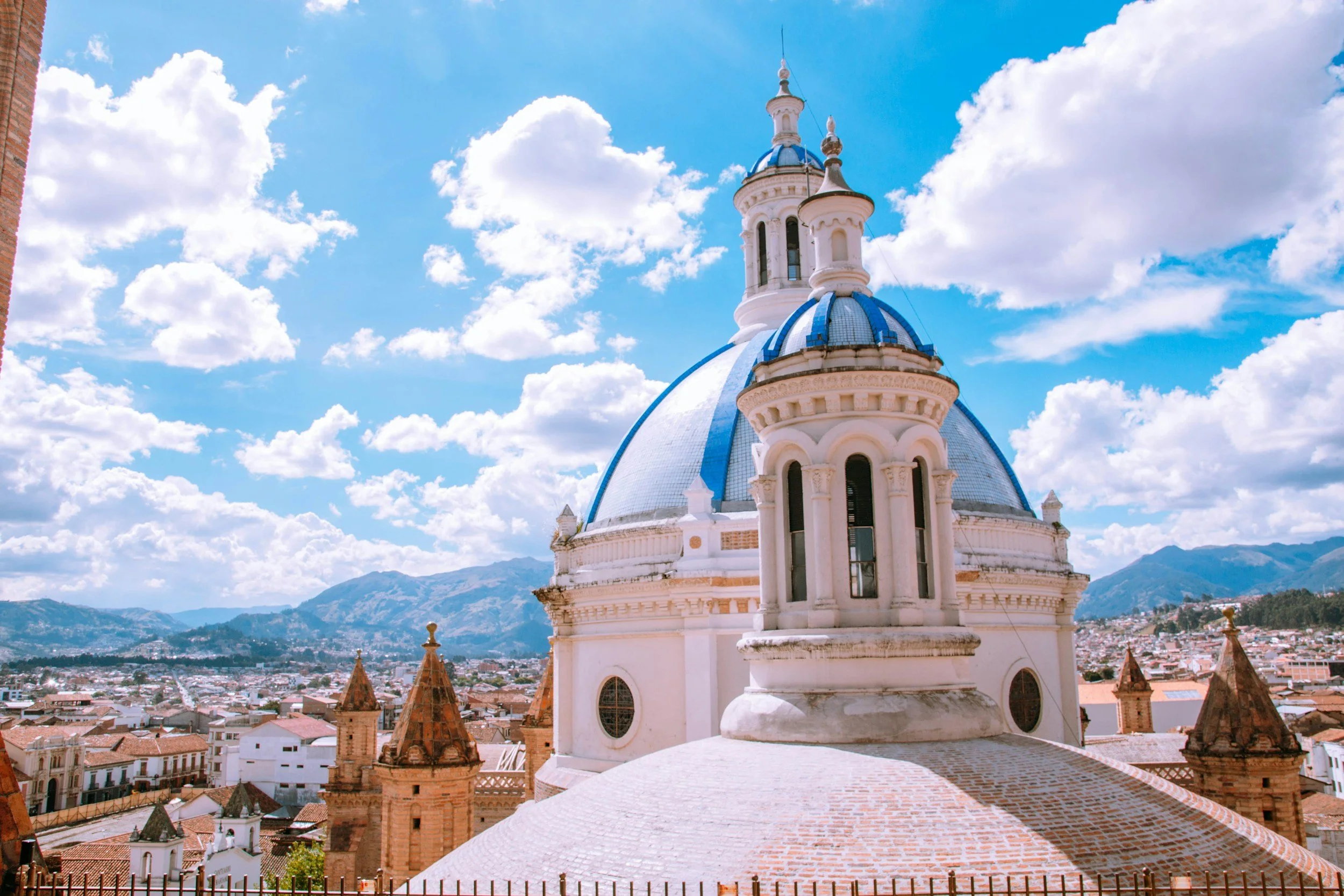 The image shows a white church with a blue domed roof and a cross on top, against a backdrop of blue sky with scattered clouds and distant mountains.