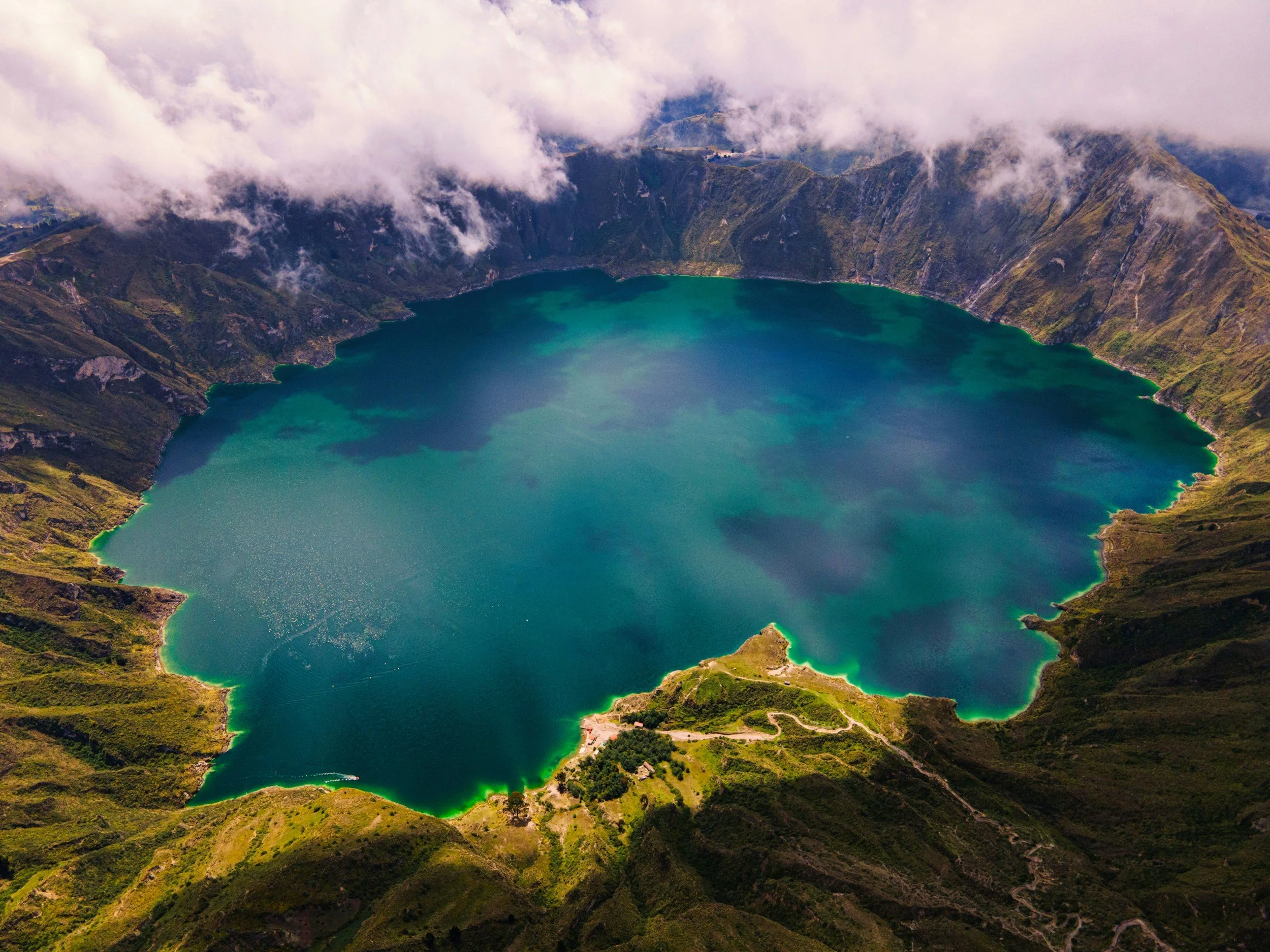 An aerial view of a volcanic crater lake surrounded by green hills and clouds in the sky.