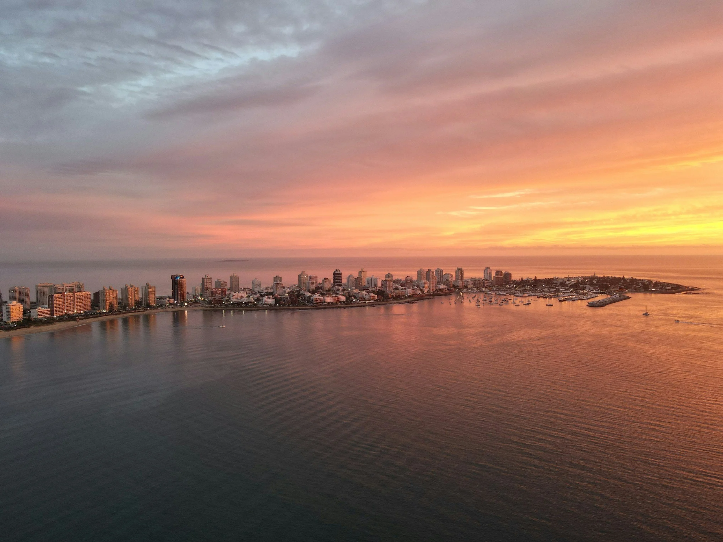 Aerial view of a city skyline near the ocean during sunset, with high-rise buildings along the shoreline, boats in the water, and a colorful sky with shades of pink, orange, and purple.