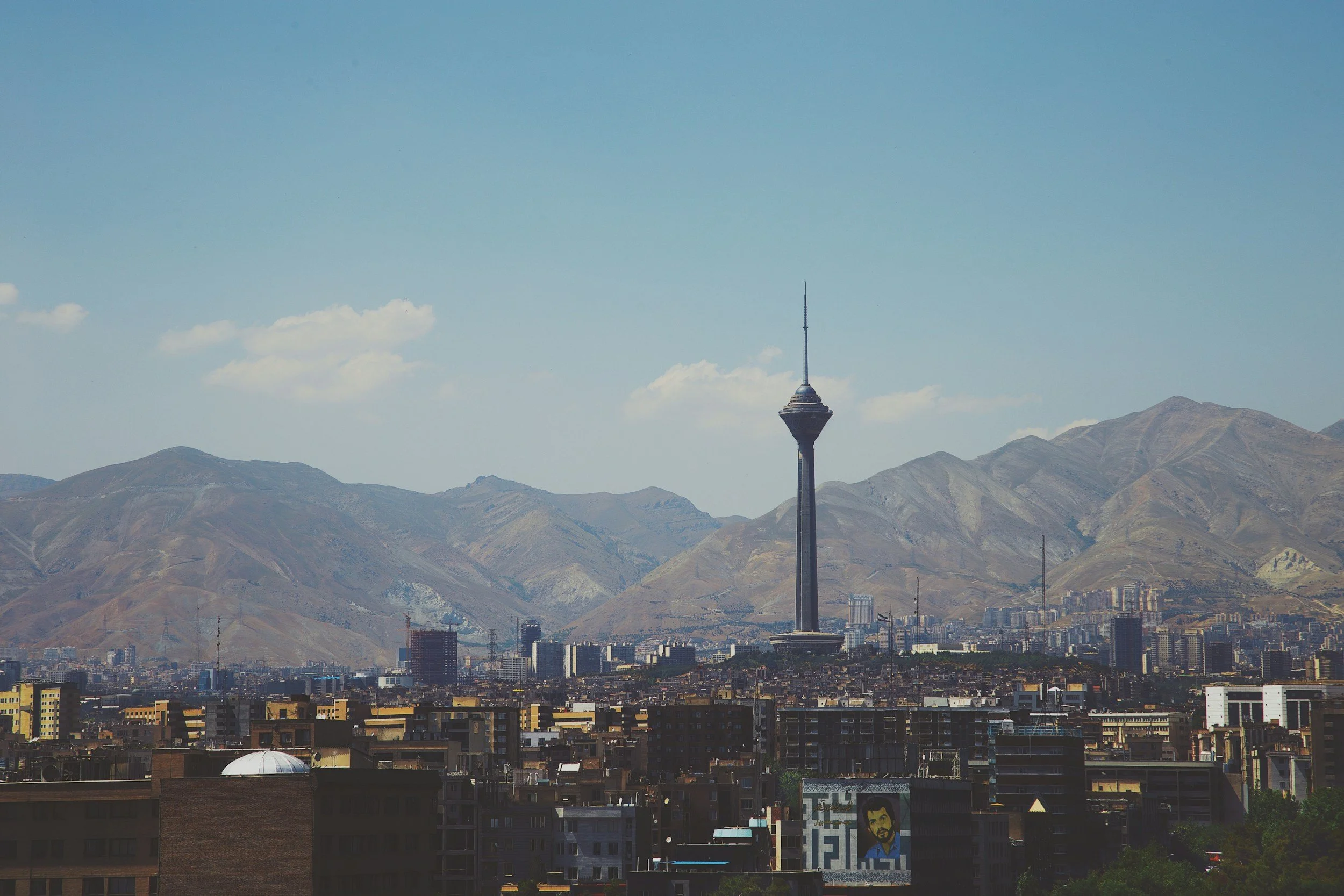 City skyline with prominent TV tower and mountain range in the background under a blue sky.