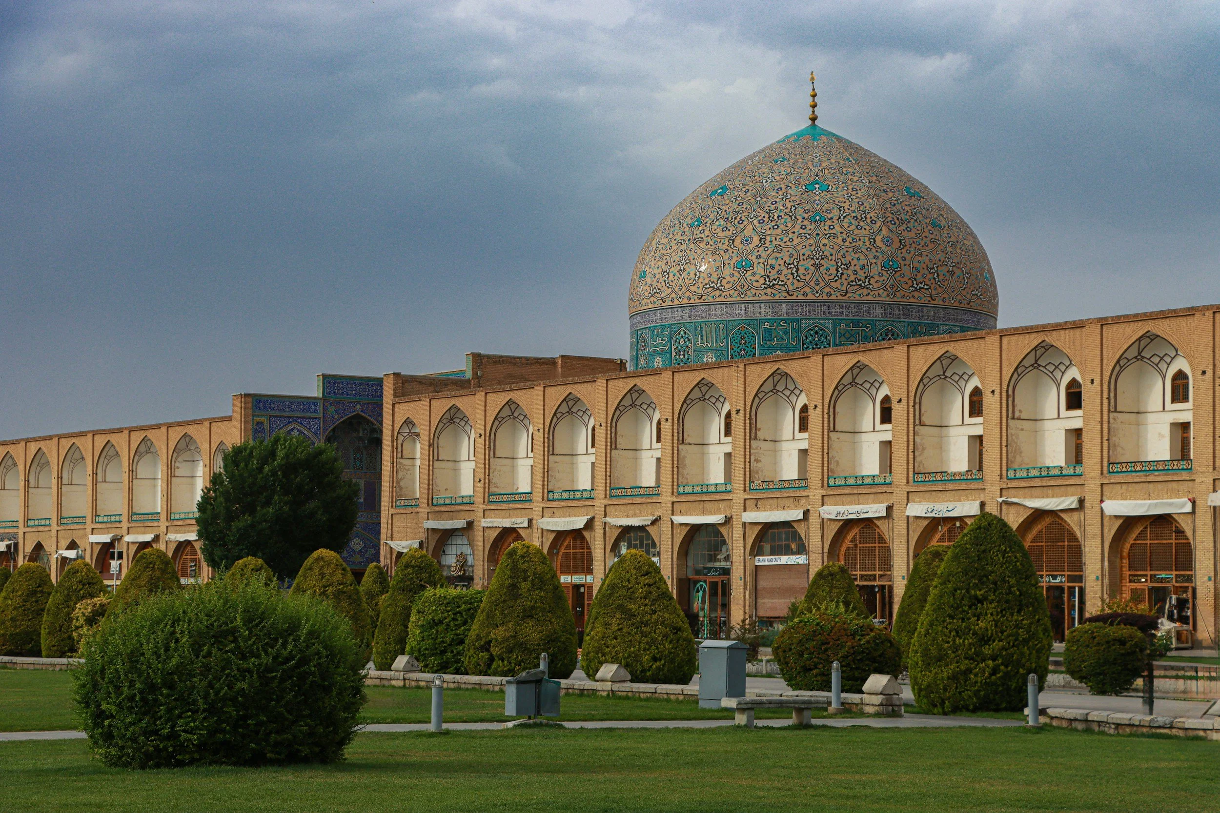 View of a historical Islamic building with a large, decorated blue and gold dome and a series of arched windows and doors, surrounded by neatly trimmed bushes and a green lawn.