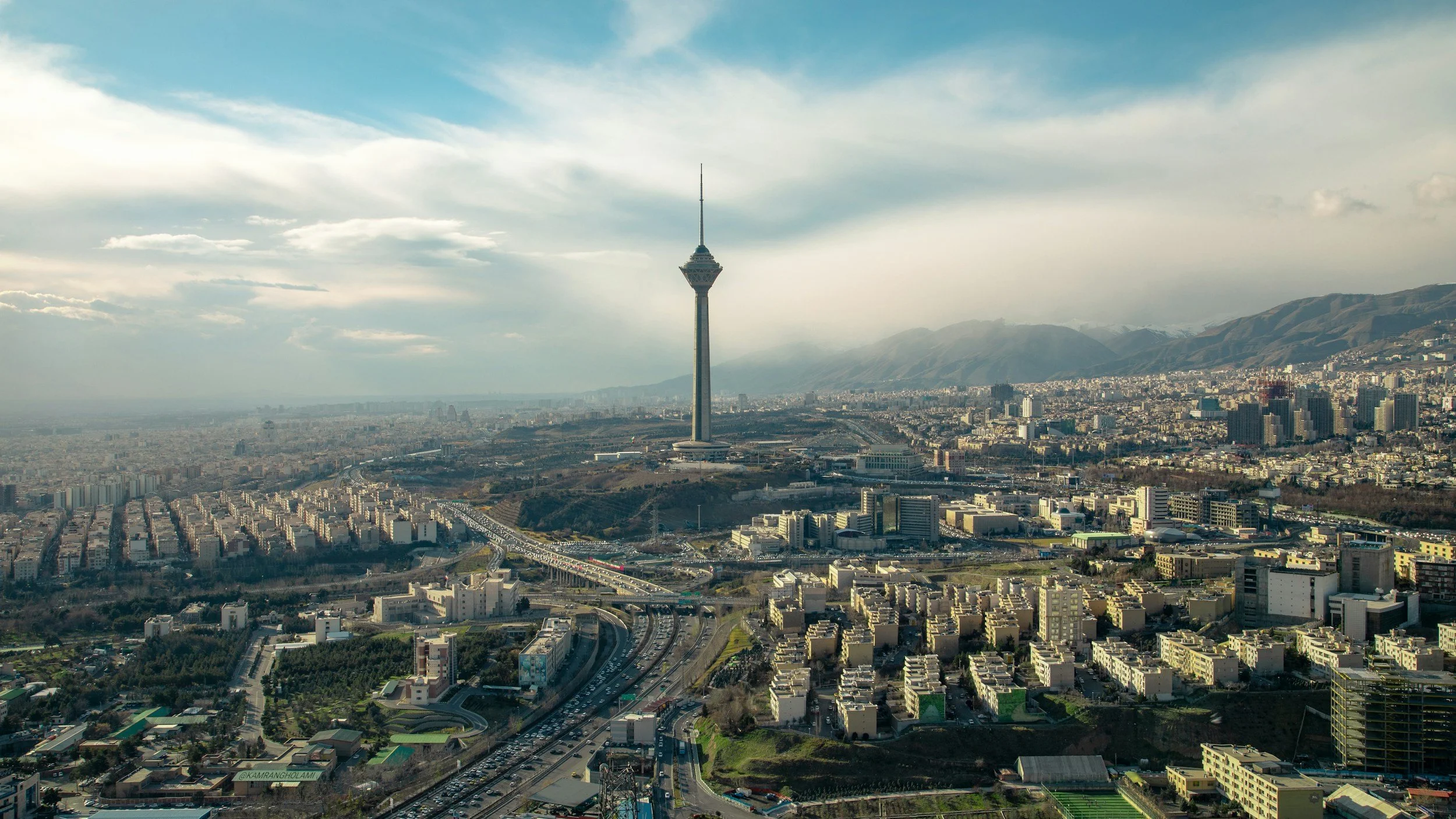 A cityscape with a tall telecommunications tower on a hill, surrounded by urban buildings, roads, and mountains in the distance.