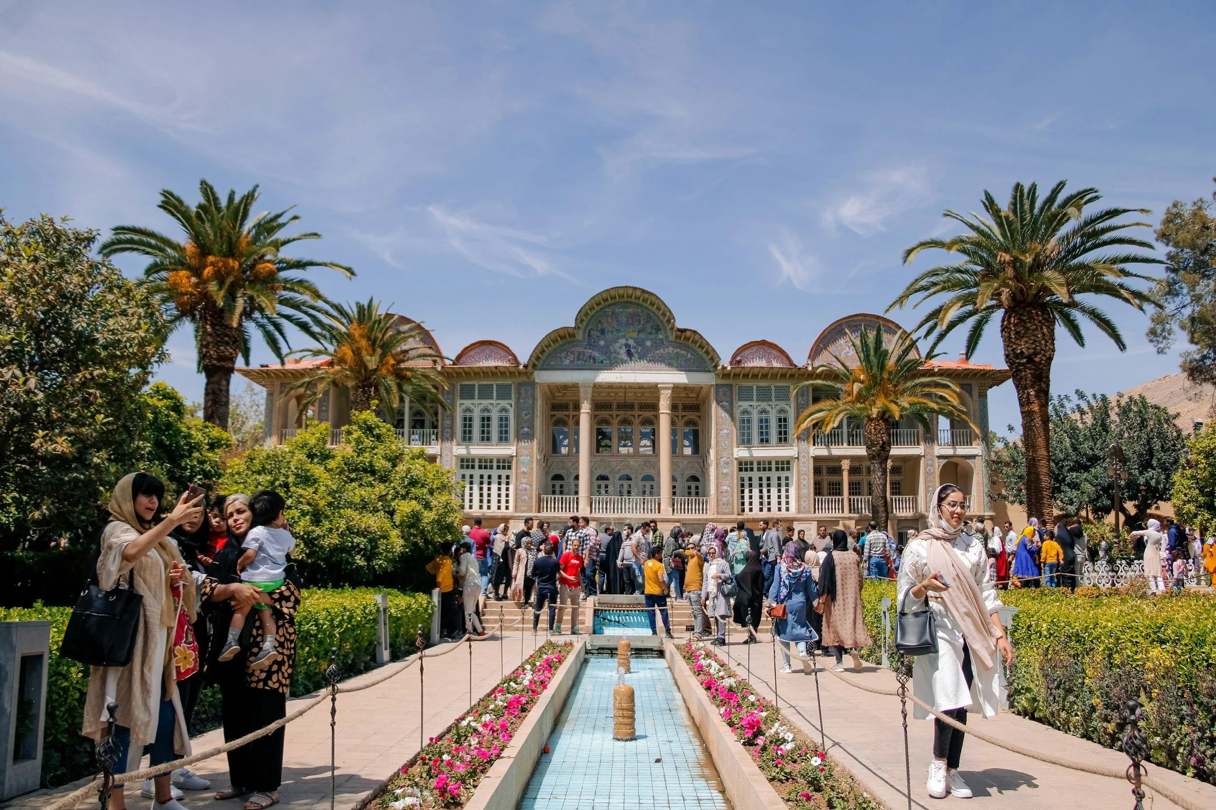 People gathered in front of the ornate, historic building with colorful mosaic details, surrounded by lush greenery and tall palm trees under a blue sky.