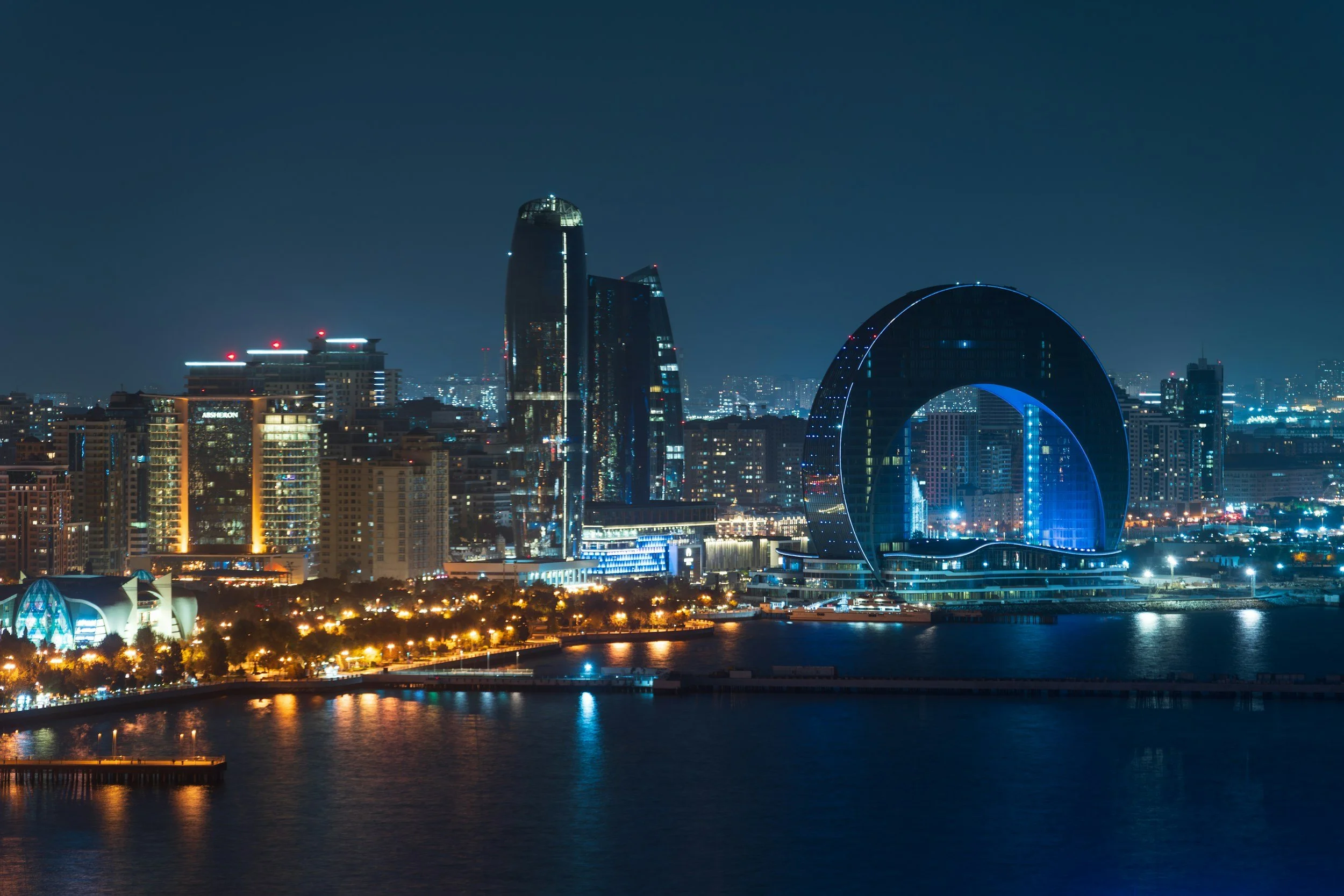 Nighttime city skyline with illuminated skyscrapers and a body of water in the foreground.