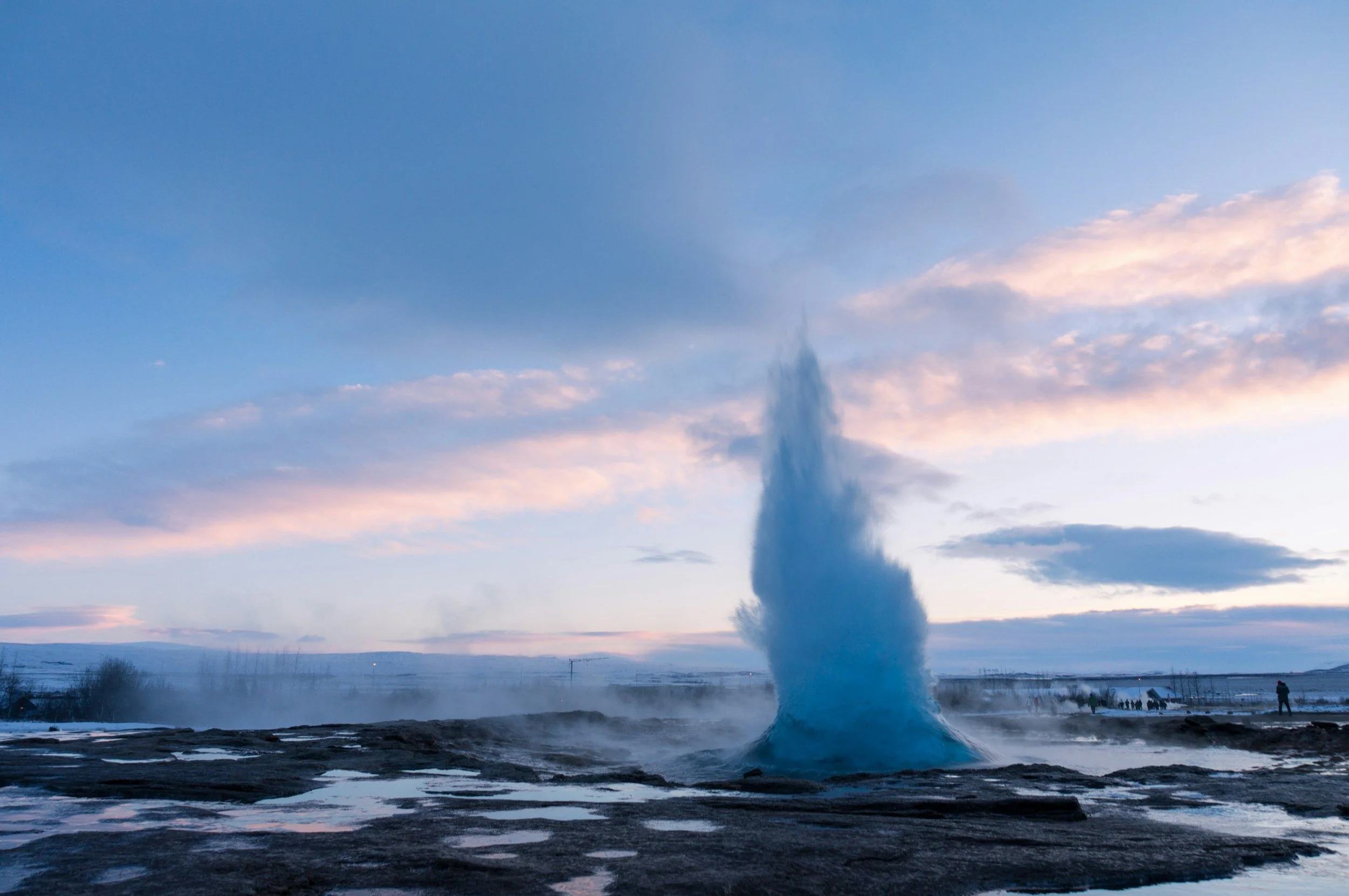 A geyser erupting and shooting water into the air with a cloudy sky at dusk in the background.